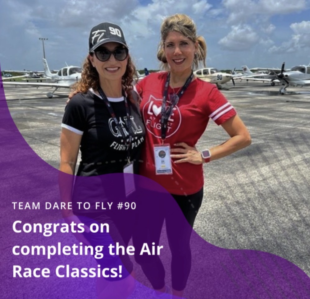 Two women smiling outdoors near small airplanes, one wearing a black shirt and cap, the other in a red shirt.