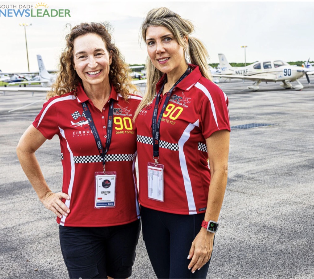 Two women in red shirts with event badges standing on tarmac with small airplanes in background.