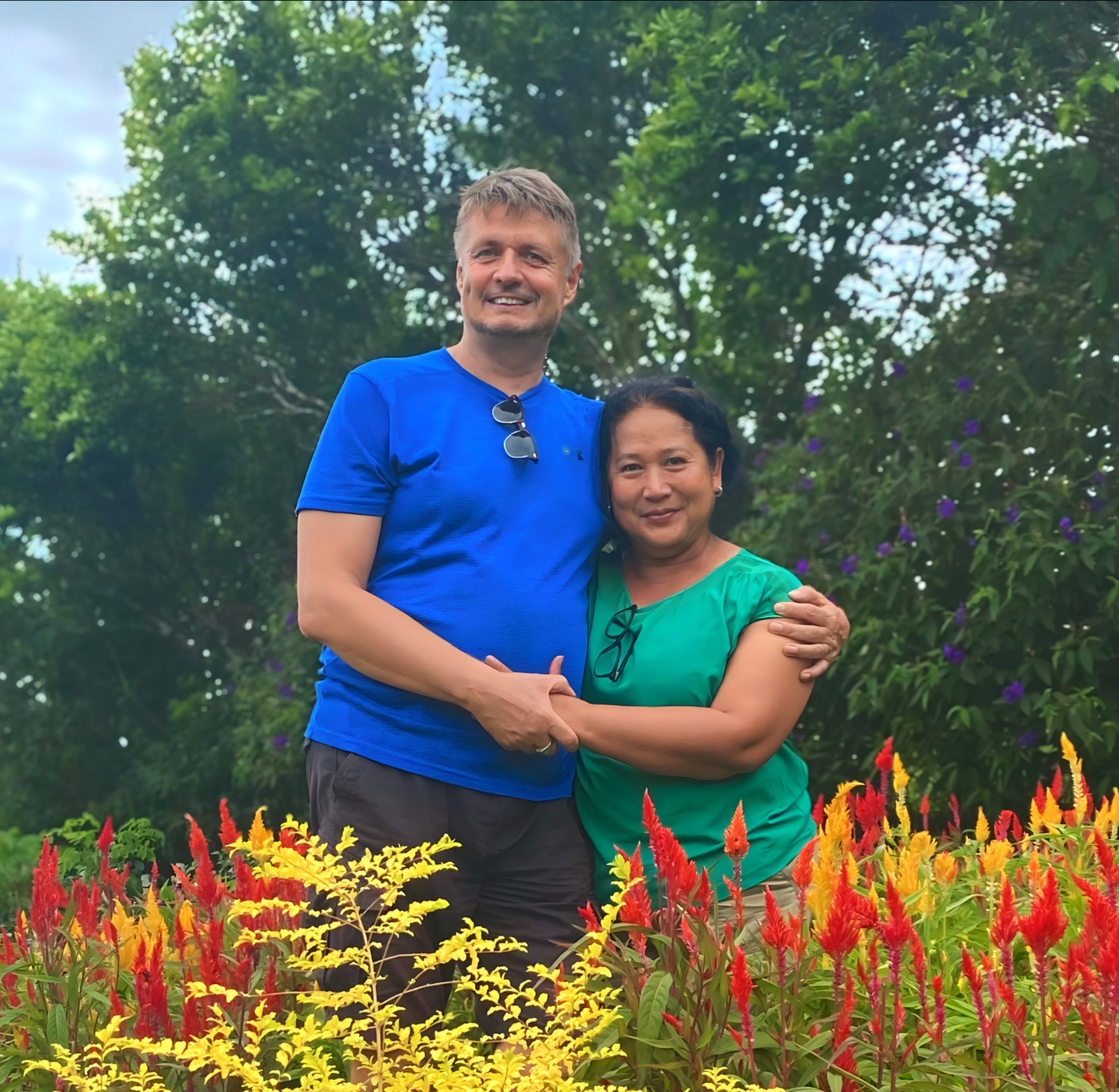 A man and woman stand together in a garden with colorful flowers and green trees in the background.