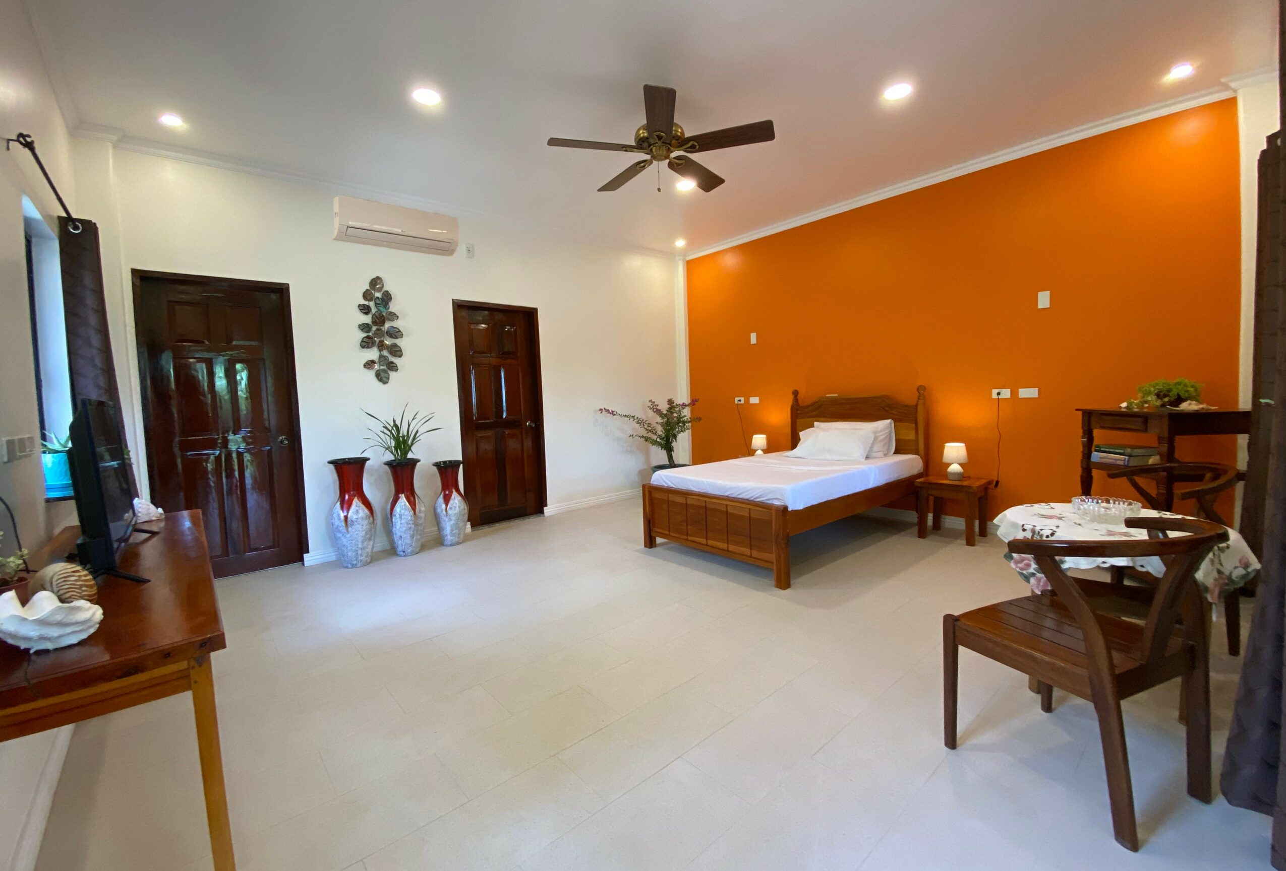 Bedroom with bed, orange accent wall, ceiling fan, wooden furniture, potted plants, and a window with curtains.