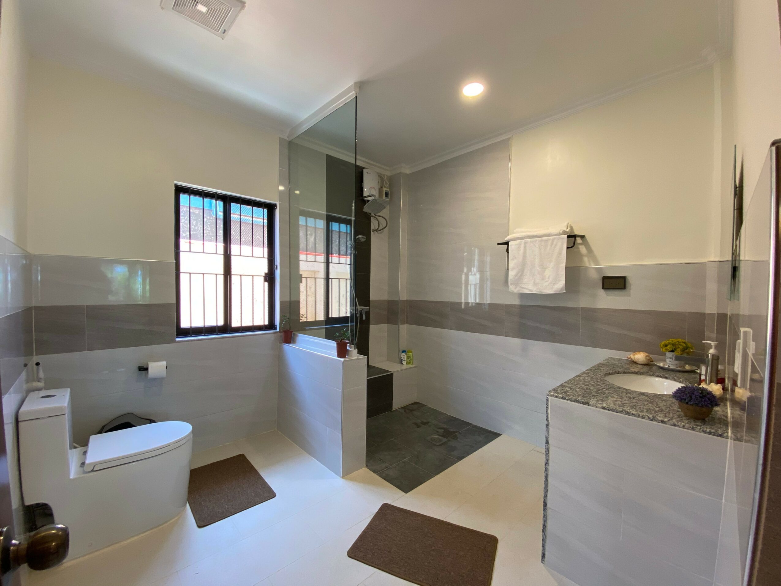 Modern bathroom with toilet, shower area, window, towel rack, and countertop with toiletries.