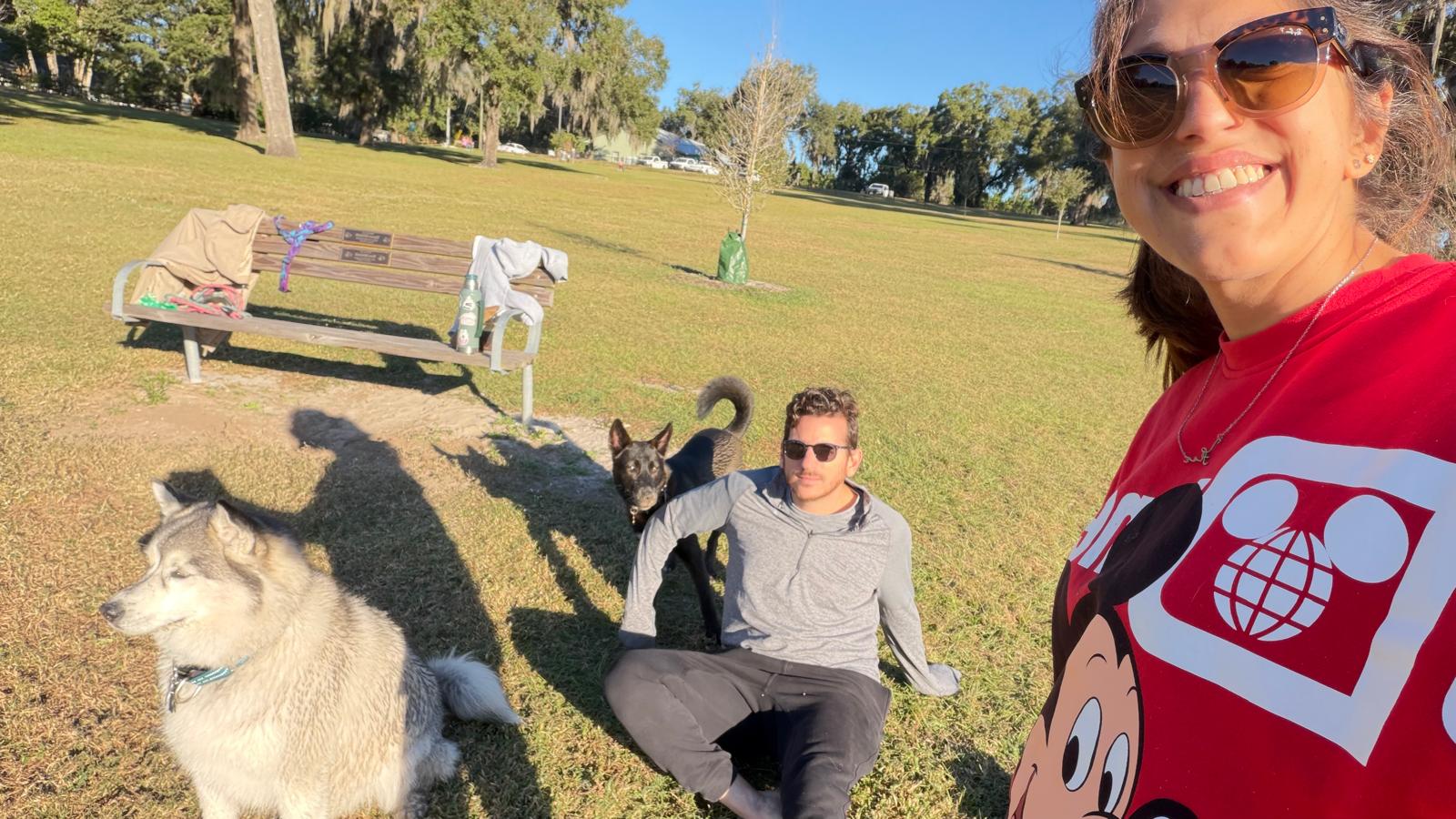 Person taking selfie outdoors with three dogs on grass, sunny day, park setting, smiling woman in red shirt, bench in background.
