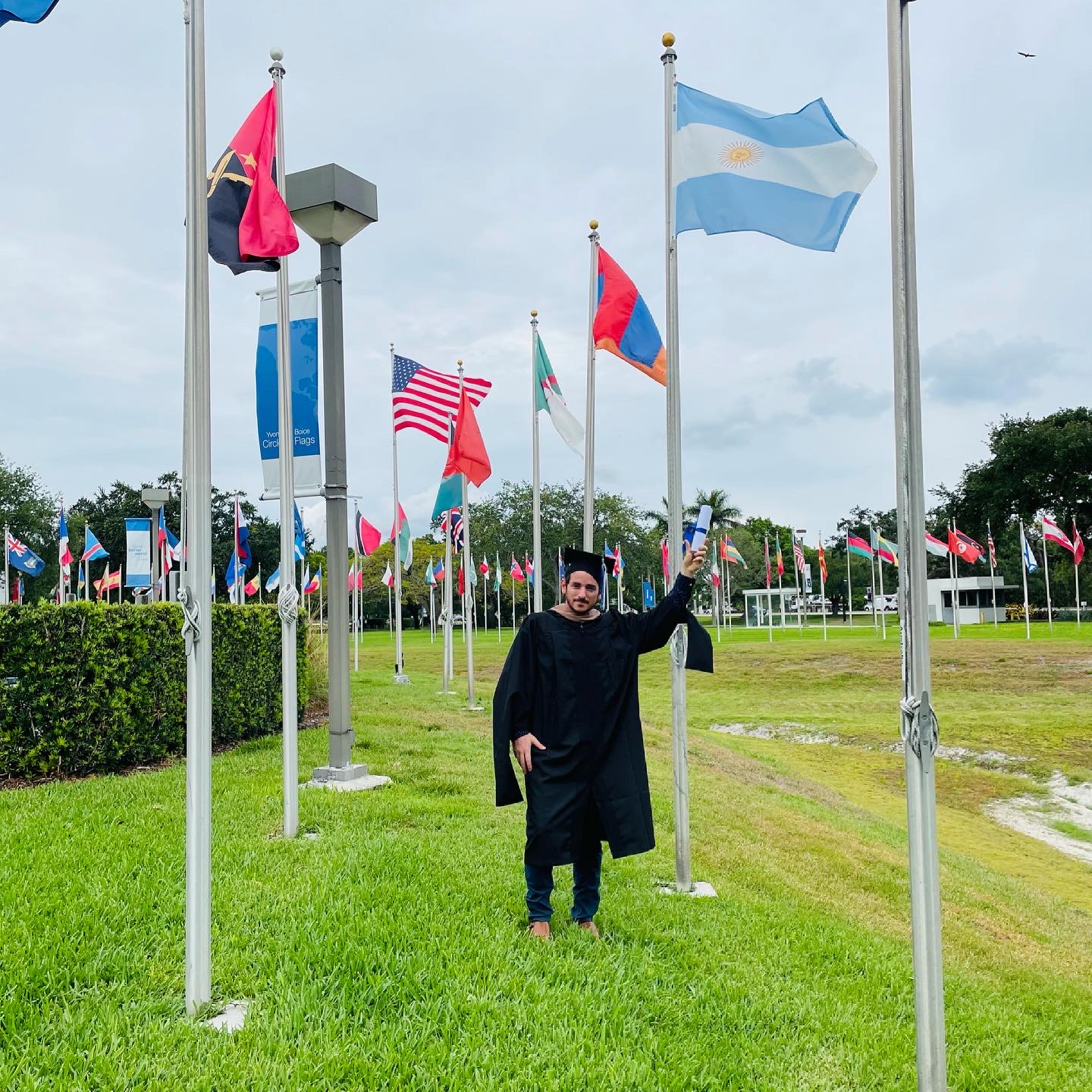 Person in graduation gown standing among flagpoles with various national flags on a grassy field.