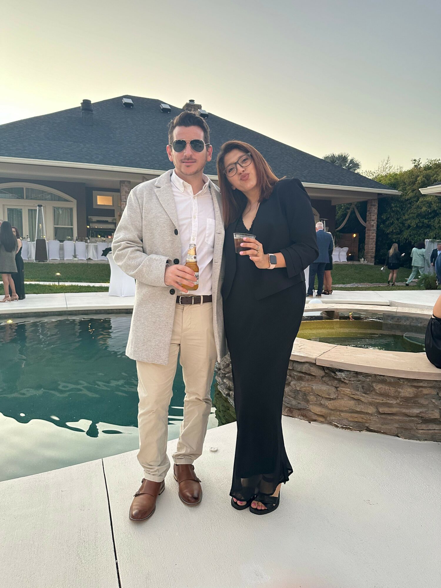 Man and woman standing by pool, holding drinks, smiling, in formal attire, outdoor event, evening sky.