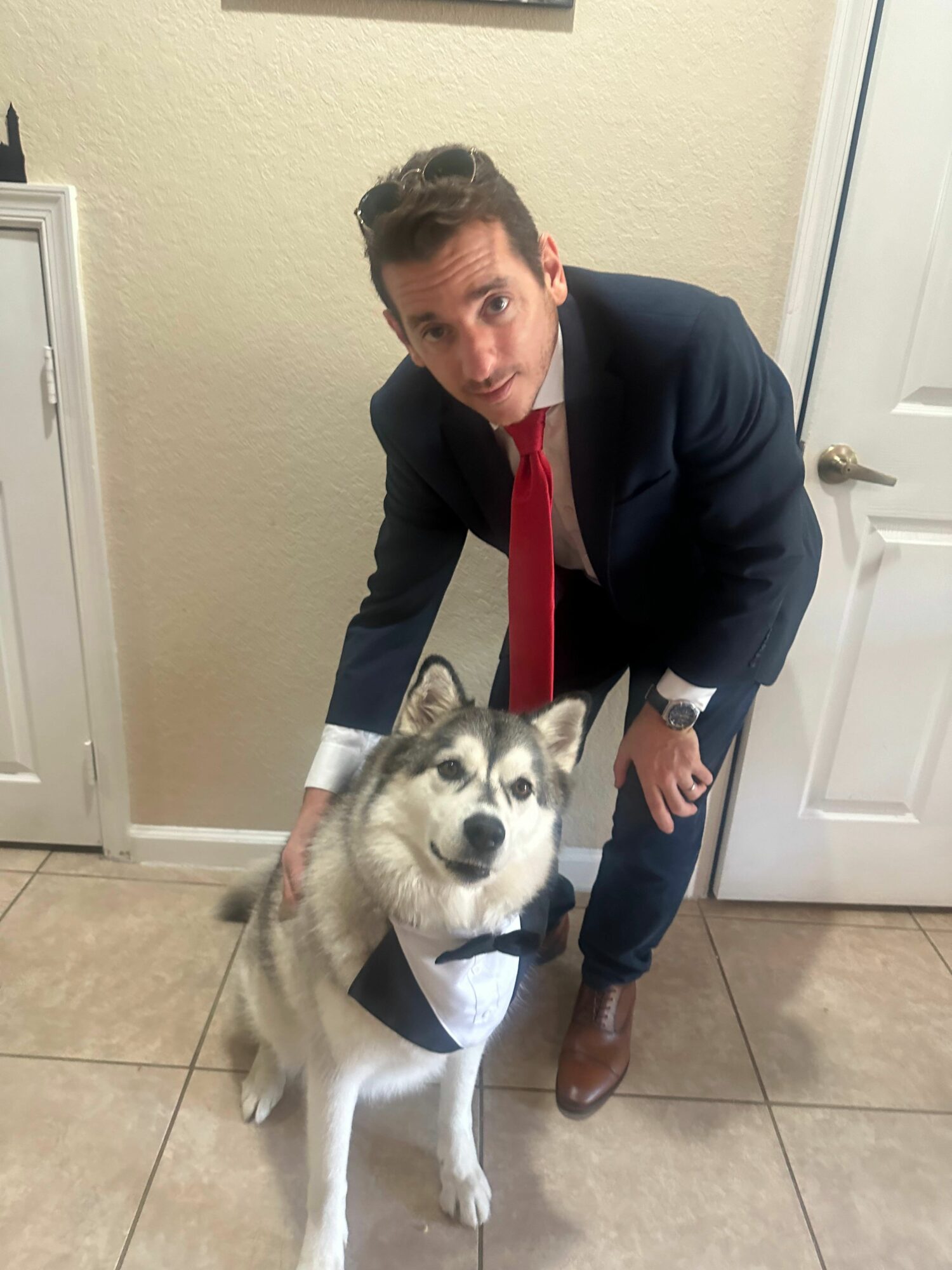 Man in suit with red tie crouching next to Siberian Husky dog inside a room.