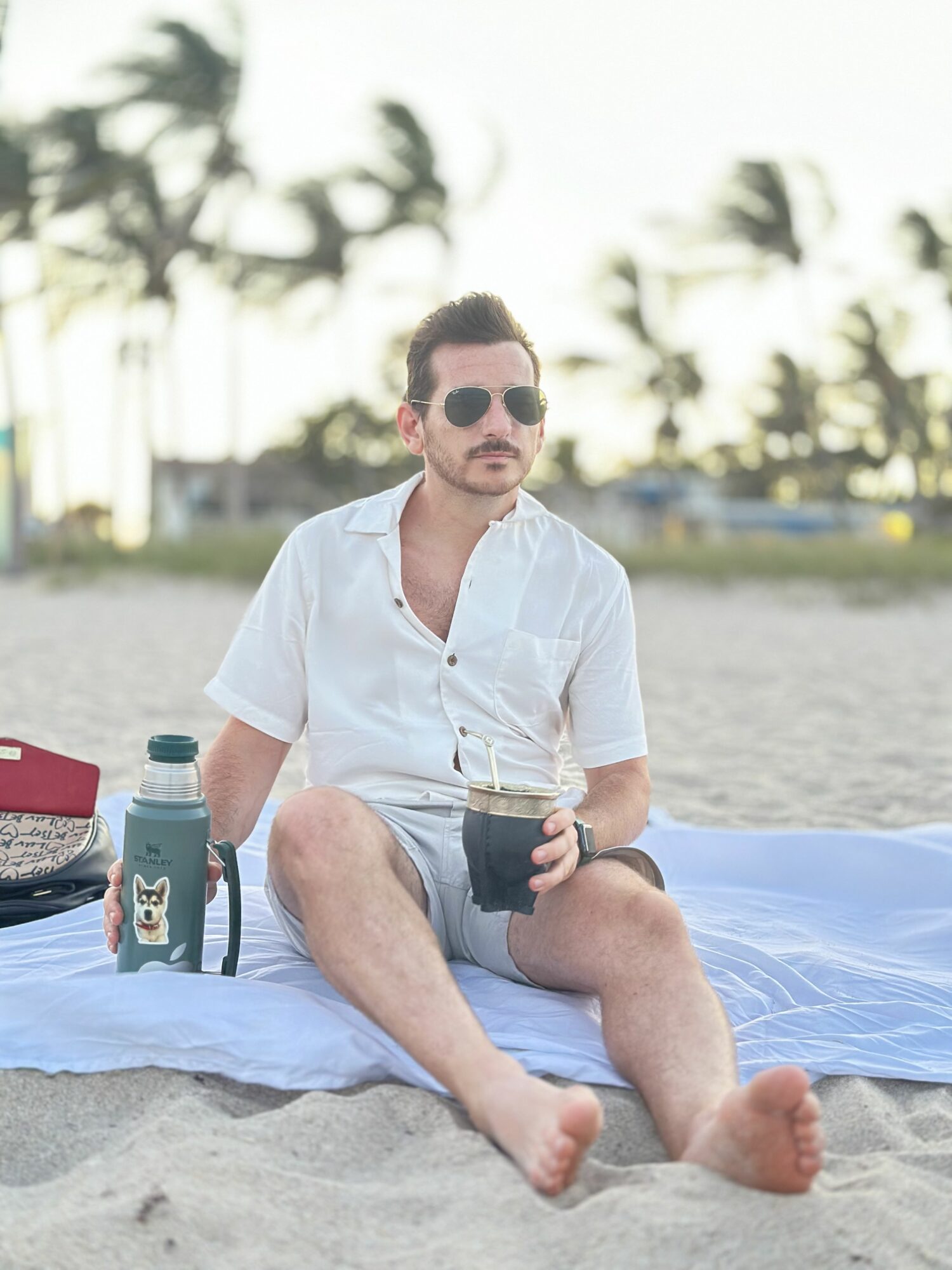 Man sitting on a blanket at the beach holding a drink, wearing sunglasses and a white shirt.