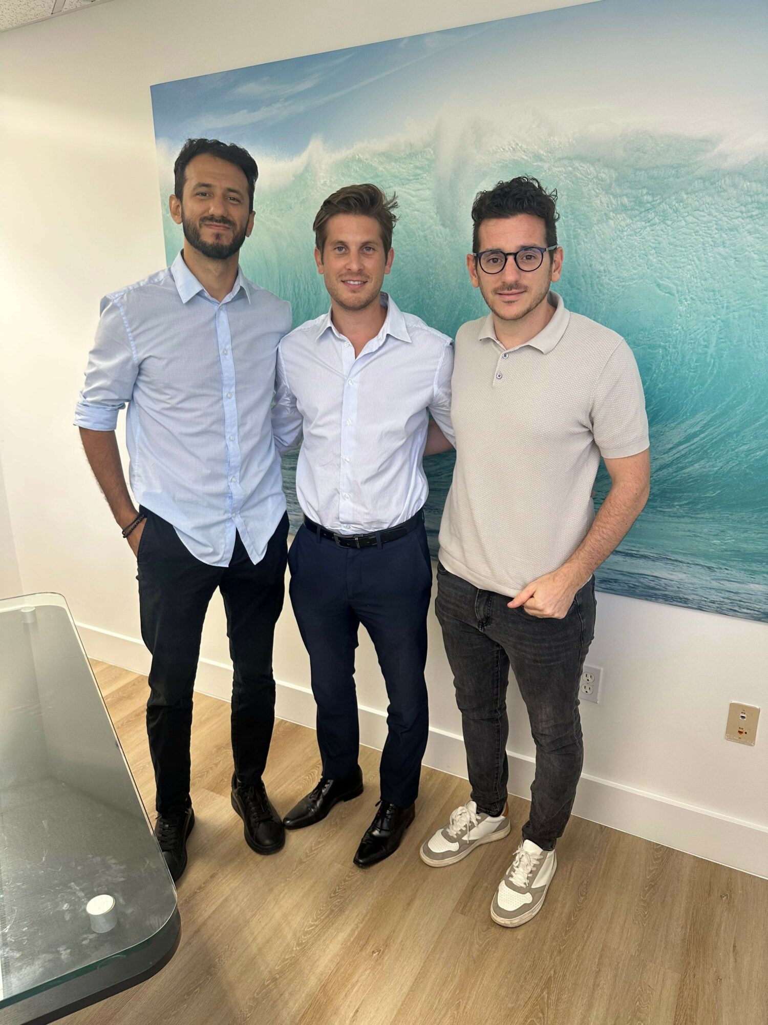 Three men standing indoors in front of a large ocean wave mural, smiling, dressed in casual and semi-formal attire.