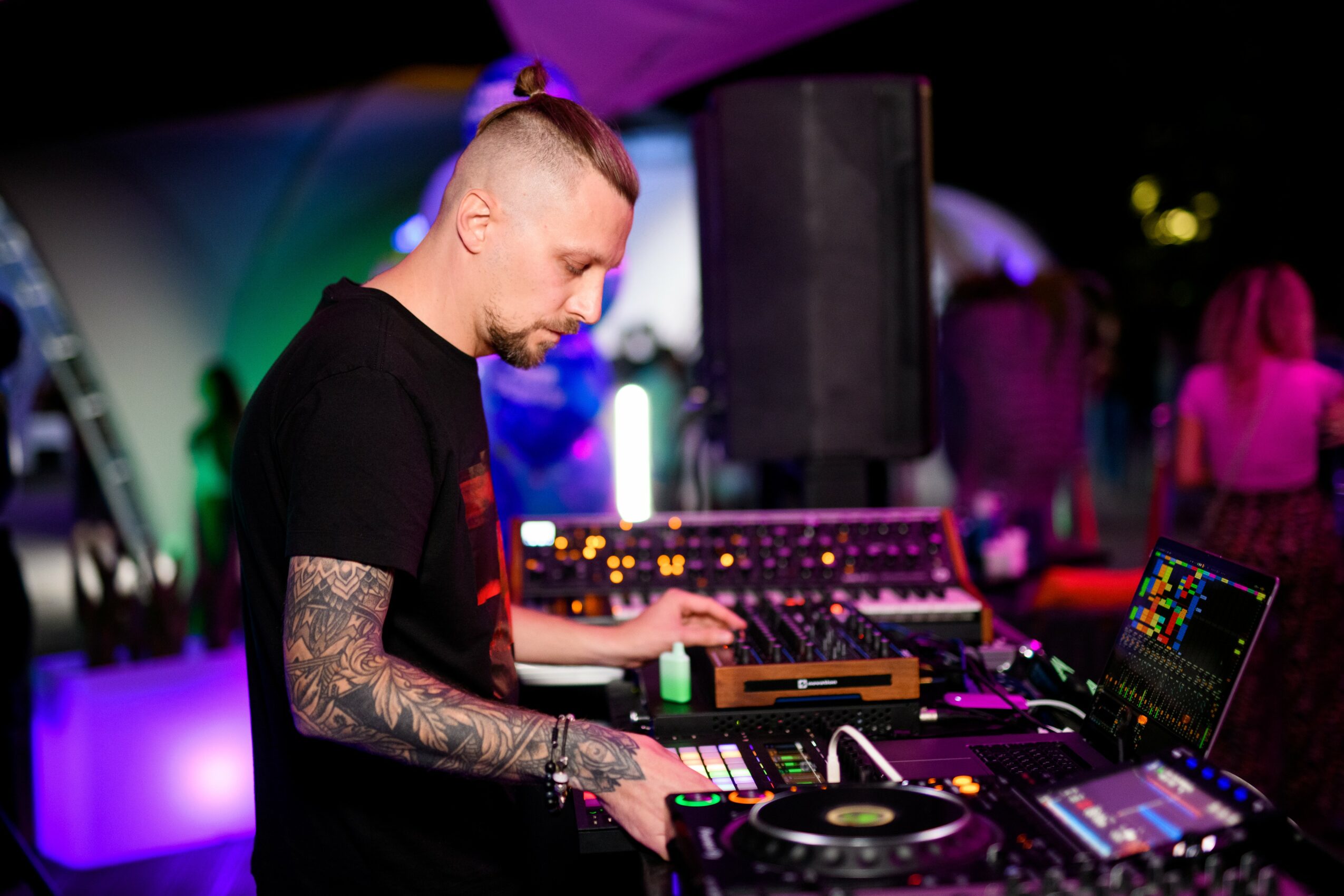DJ with tattoos on arms operating music equipment at an outdoor event at night.