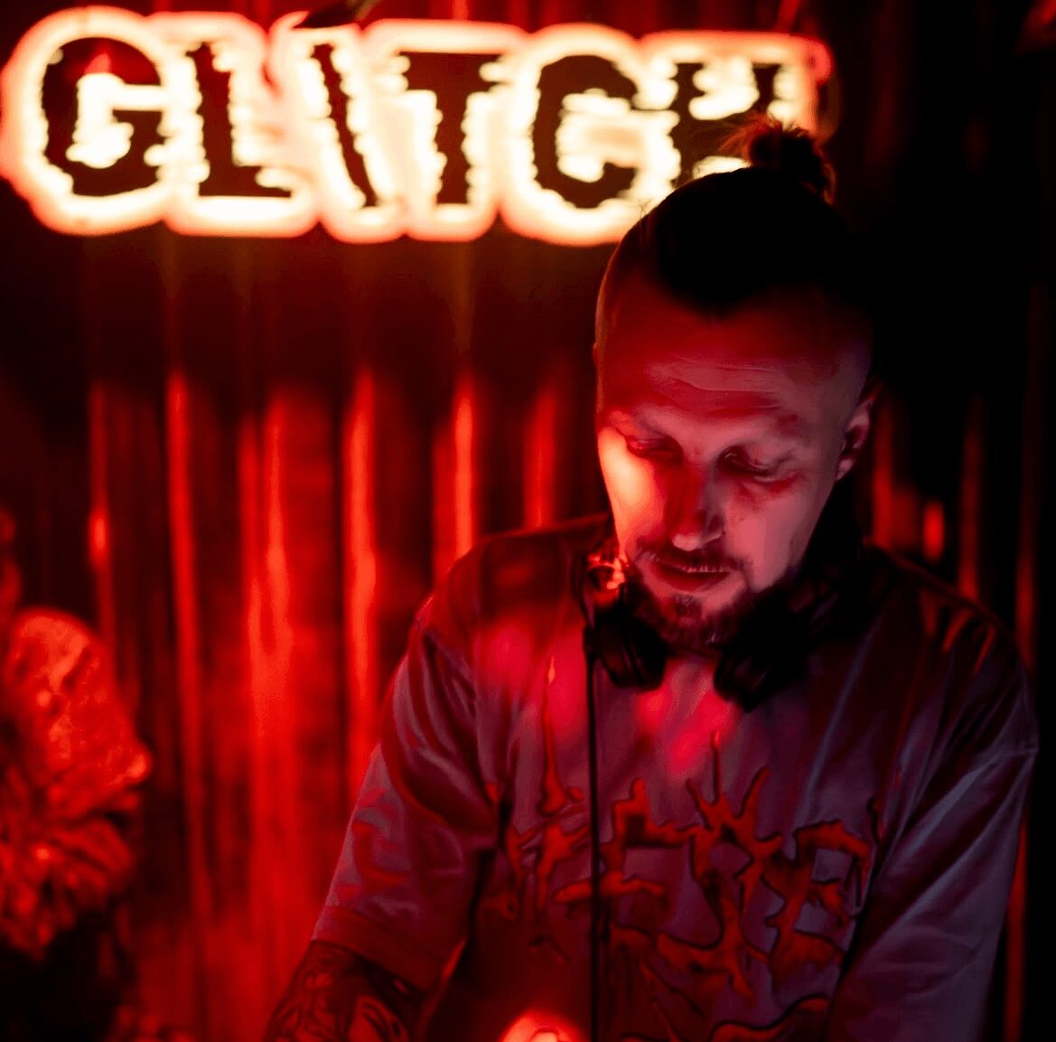 Man with headphones looking down in a dimly lit room with a red neon sign behind him.