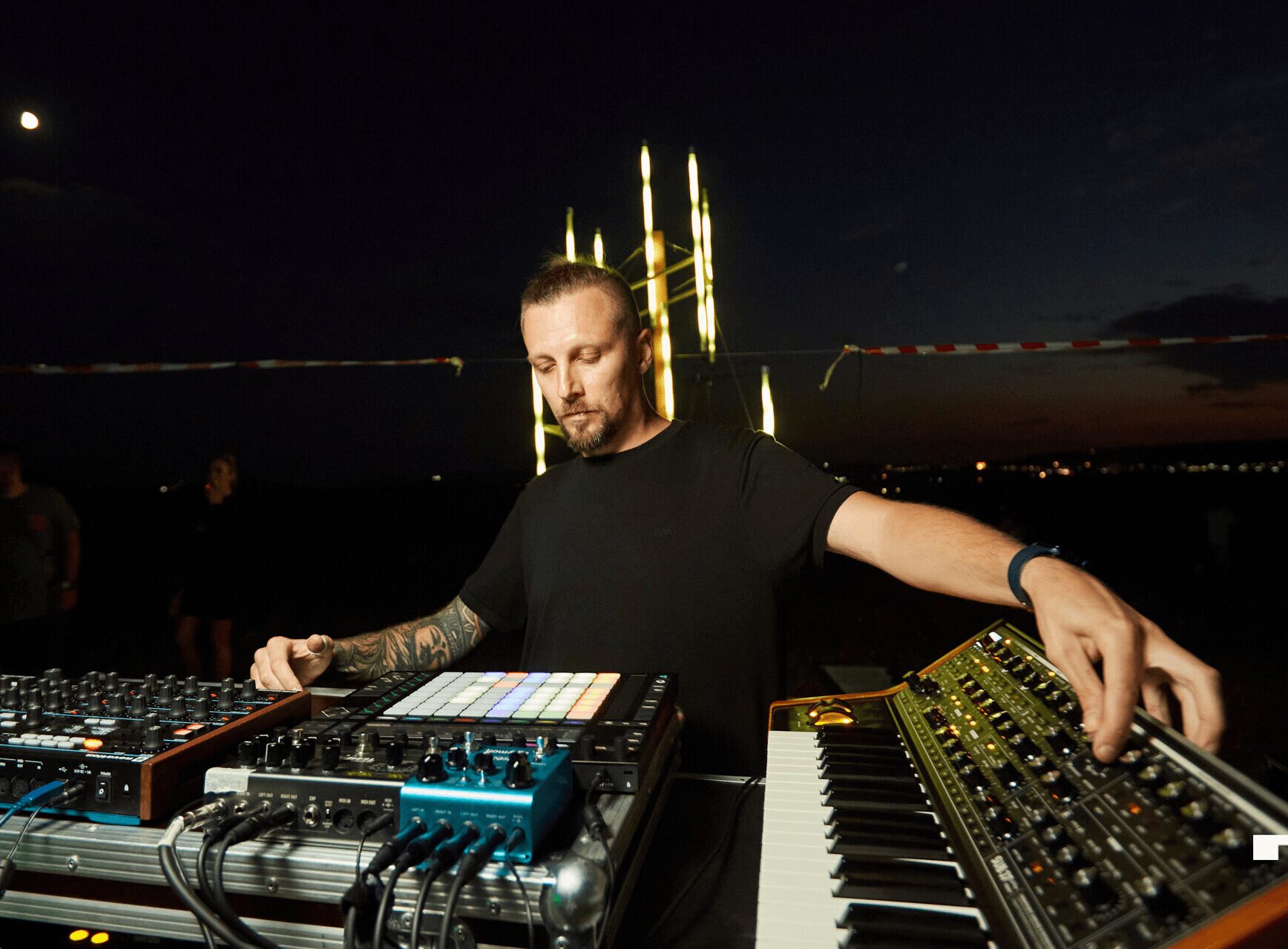DJ with equipment outdoors at night, city skyline and illuminated structures in background.