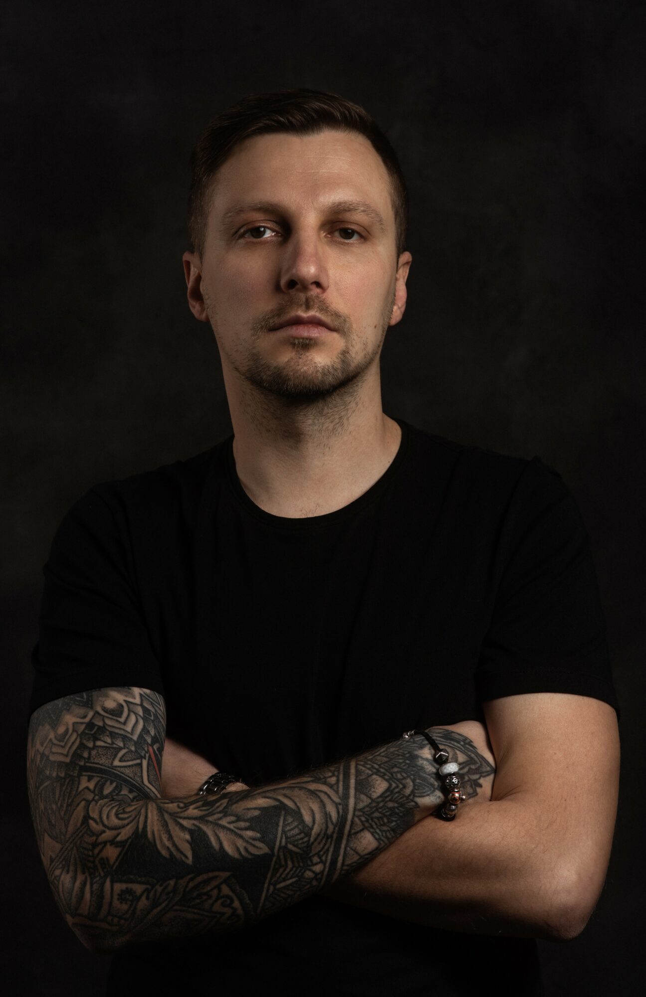 Young man with short dark hair and tattoos on his left arm, wearing a black shirt, standing against a dark background.
