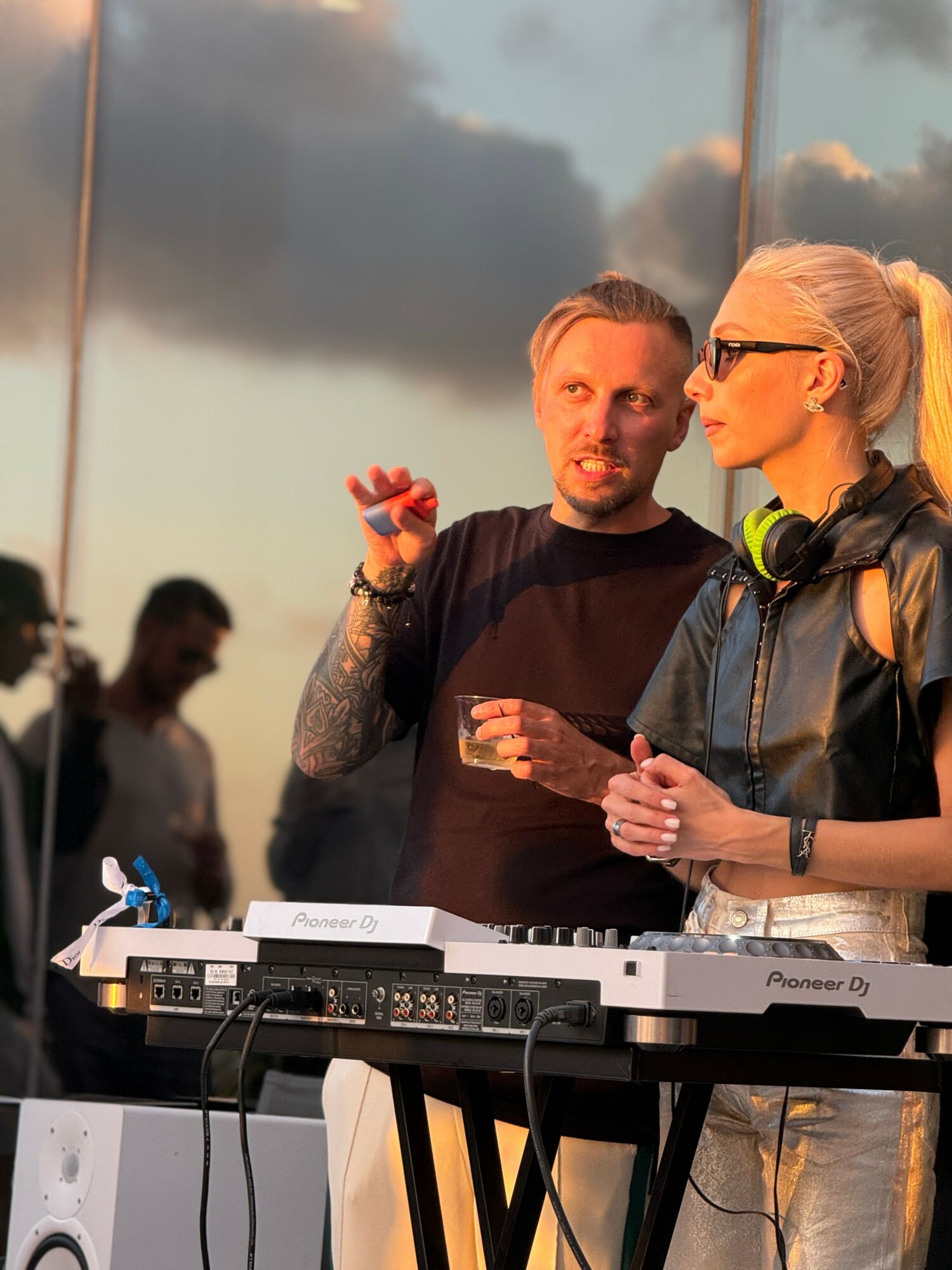 Two people, a man and a woman, stand near DJ equipment outdoors during sunset, engaged in conversation.