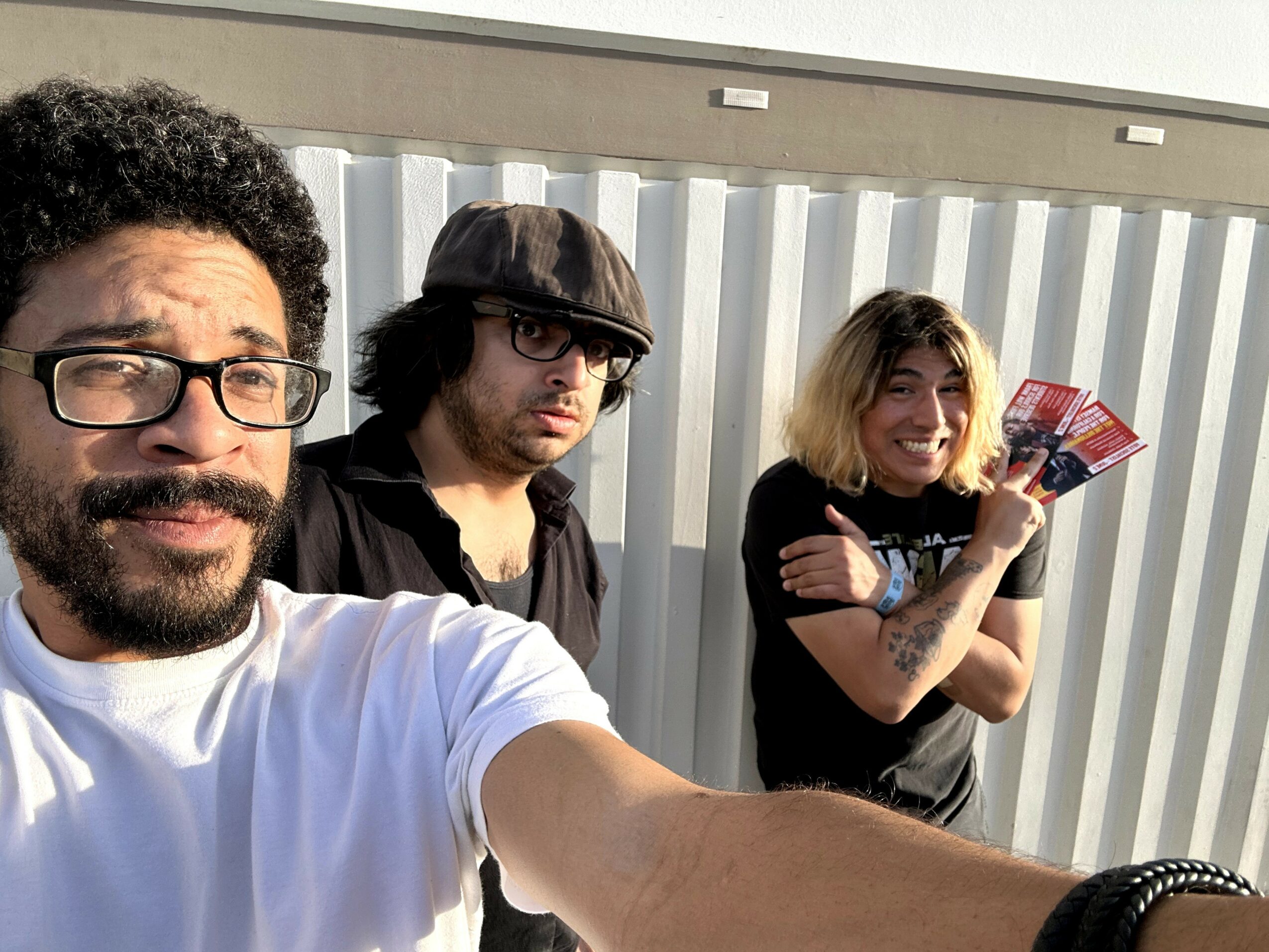 Three people standing outdoors in front of a metal fence, smiling and posing for a selfie.