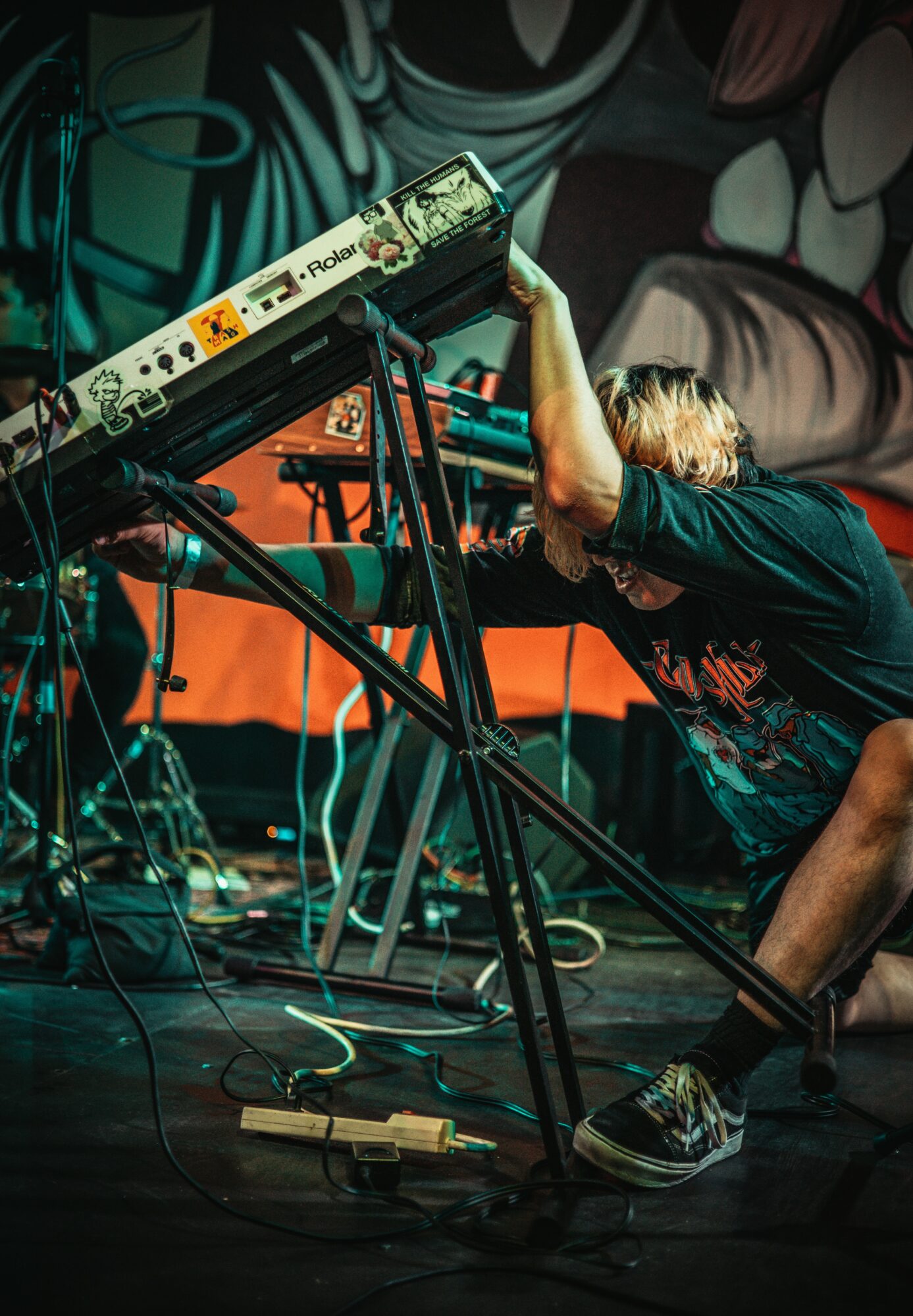 Person with blonde hair leaning over electronic equipment on stage, surrounded by cables and a pedal, in a dark setting.