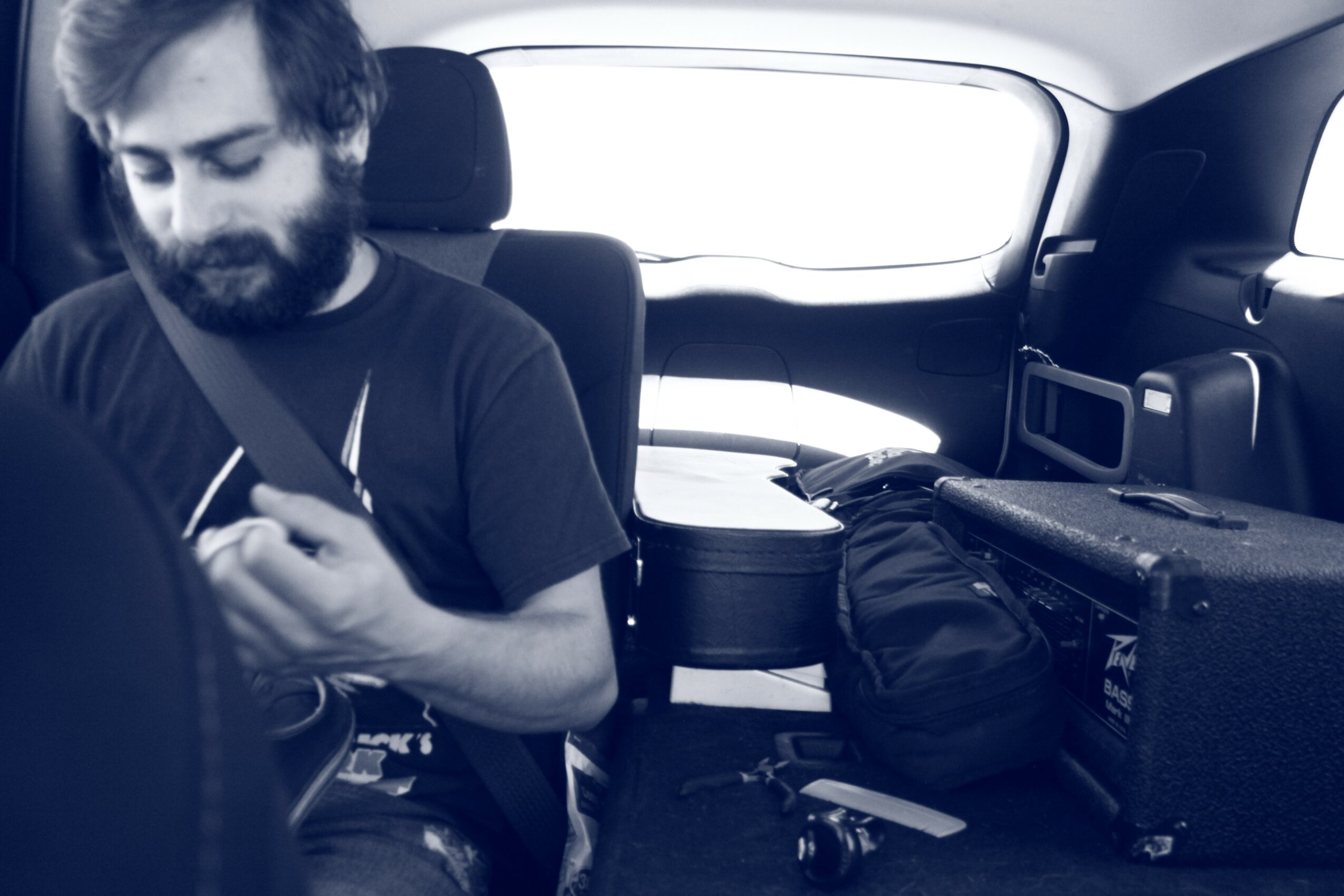 Man with beard sitting in car, looking at phone, with bags and items around him.