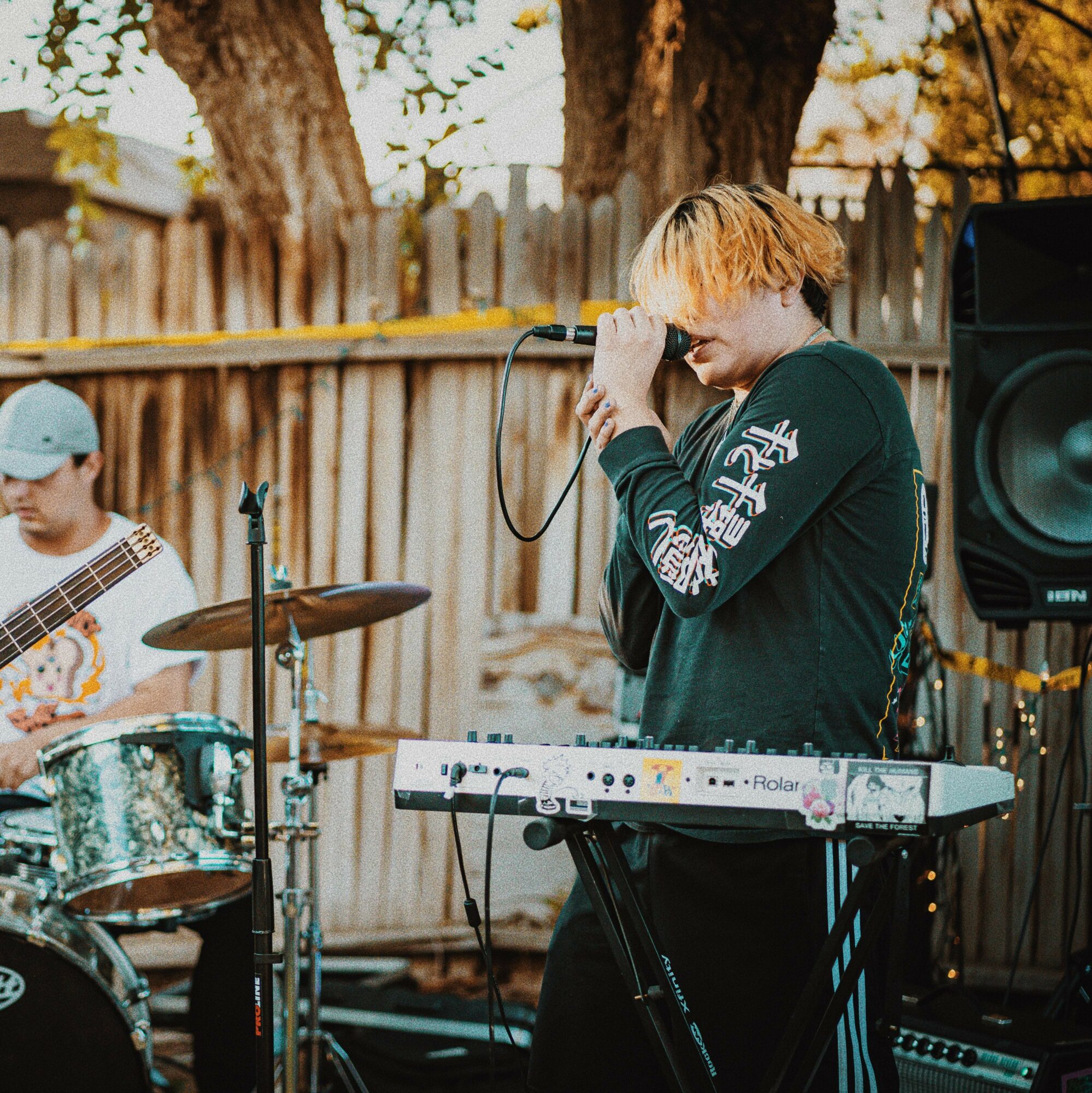 Person singing into microphone with keyboard and drum set outdoors, wooden fence and trees in background.