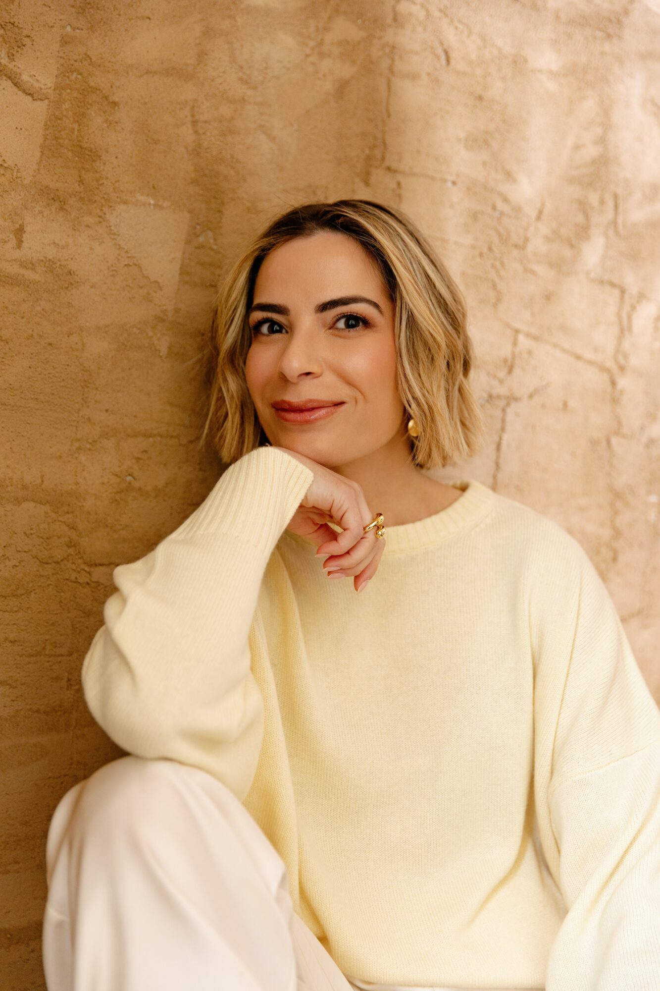 Woman with shoulder-length blonde hair wearing a cream sweater, sitting against a textured beige wall.