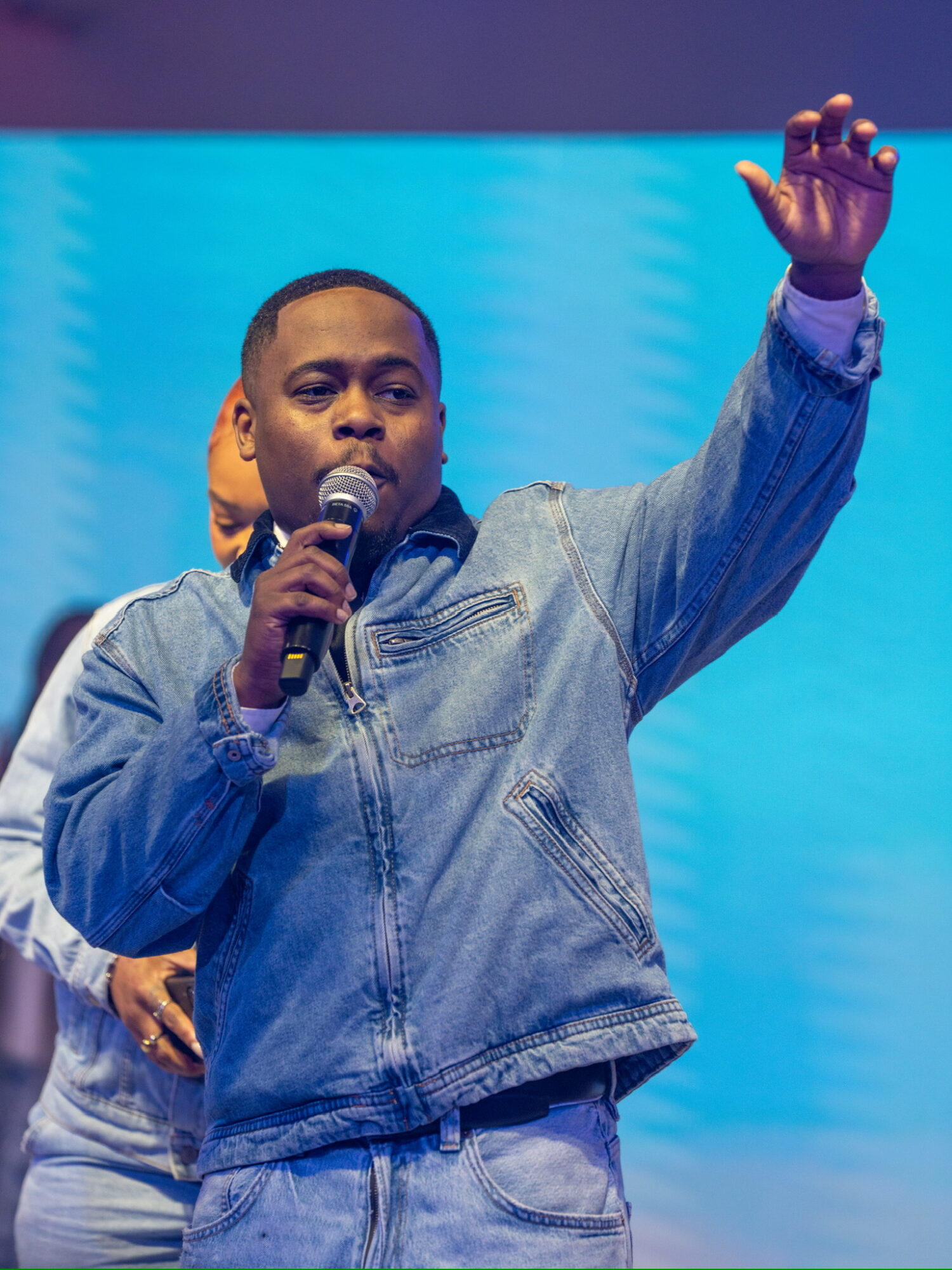 Man speaking into microphone with raised hand on stage, blue background, wearing denim jacket and jeans.