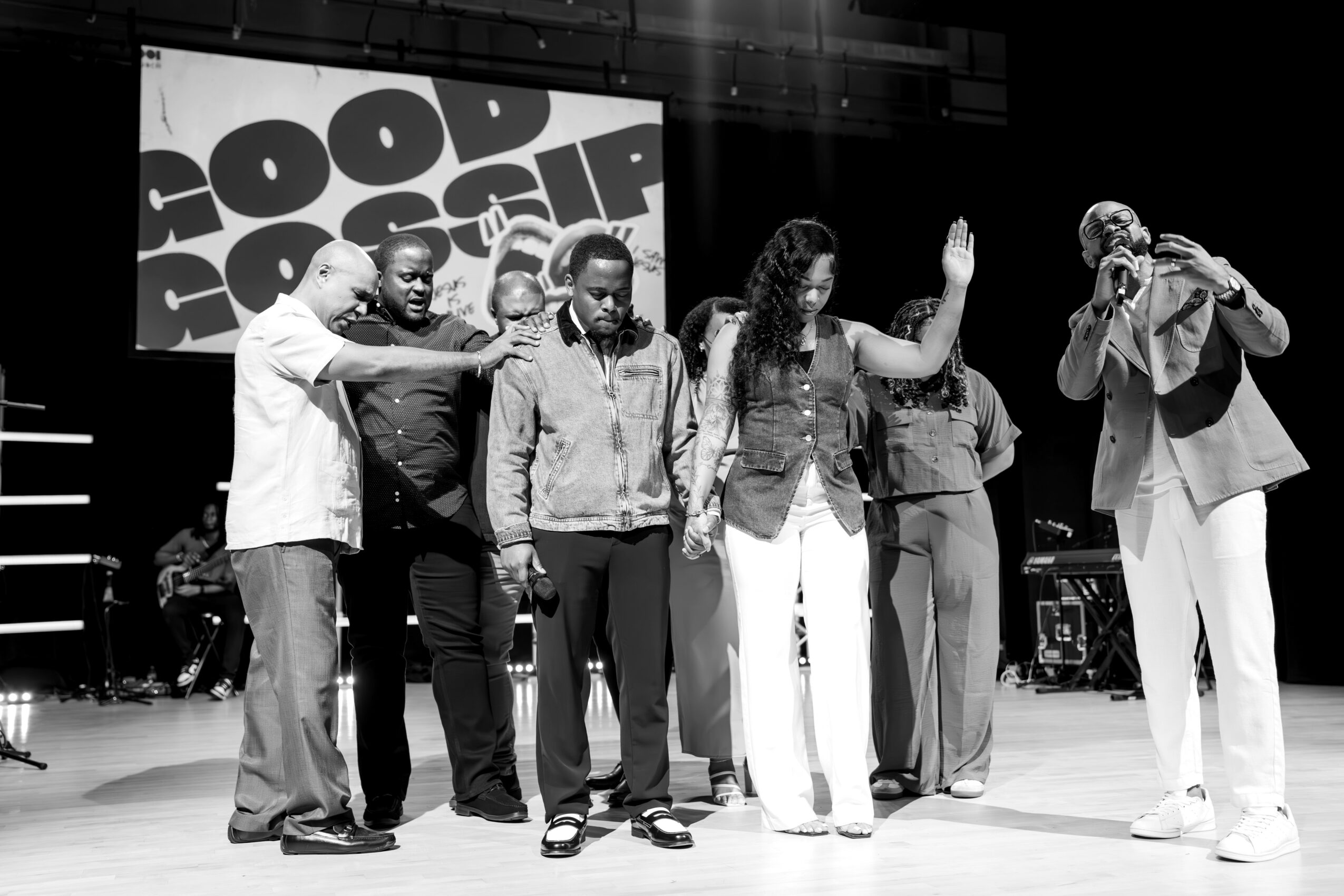 Group of seven people on stage, some with hands raised, in front of a screen with text, in black and white.