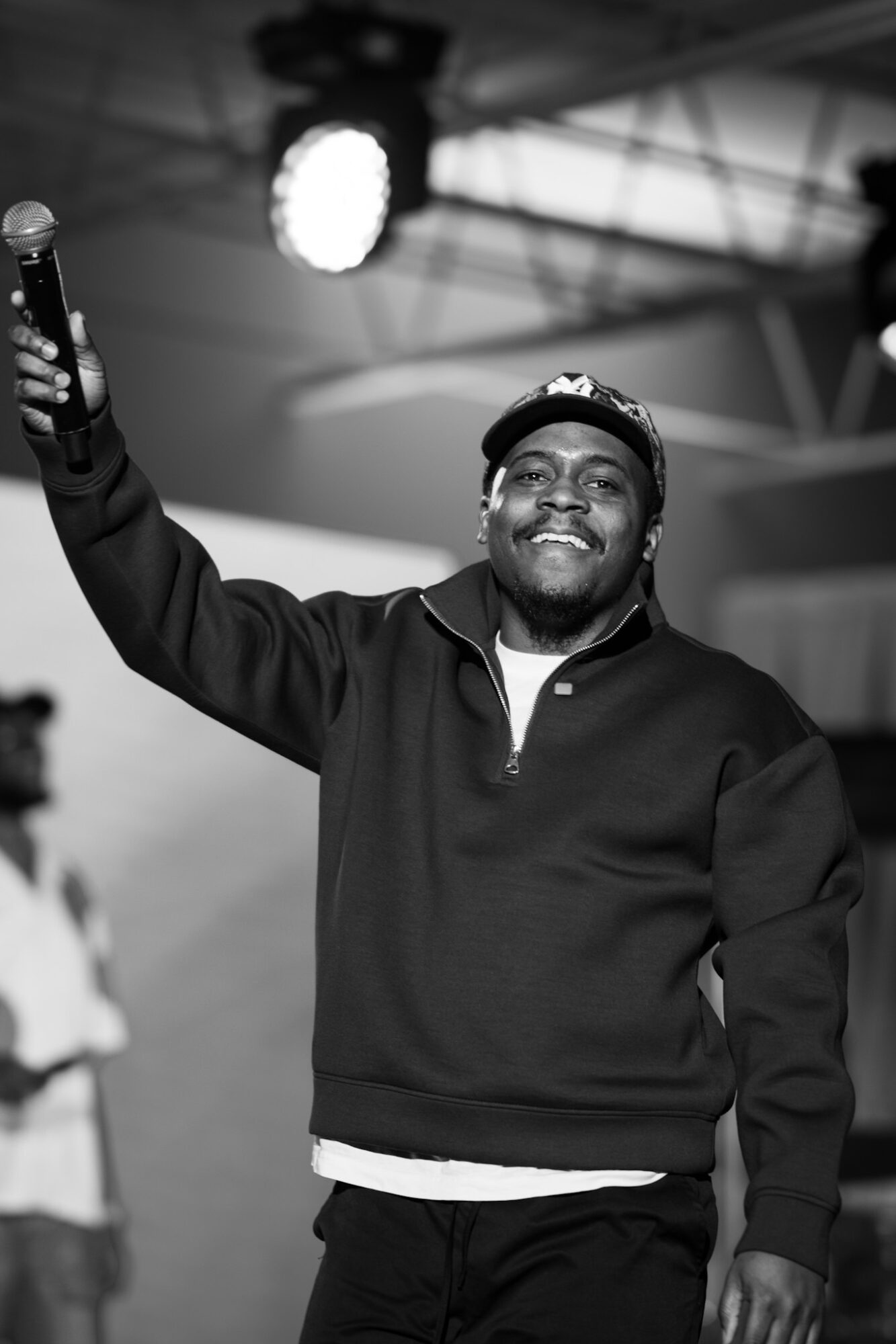 Person smiling, holding microphone, wearing cap and dark jacket, on stage with lighting and structure overhead.