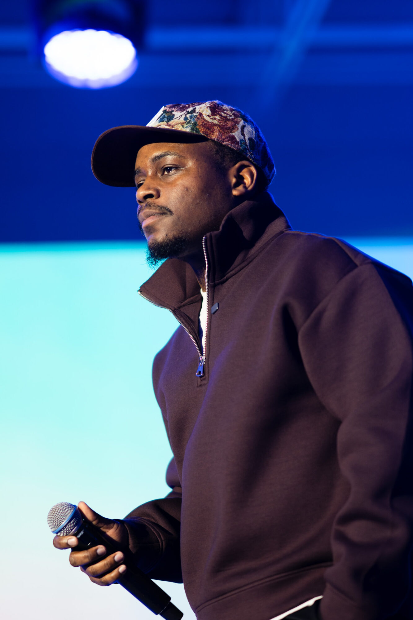 Young man wearing a patterned cap and dark jacket, holding a microphone, standing on stage with blue background.