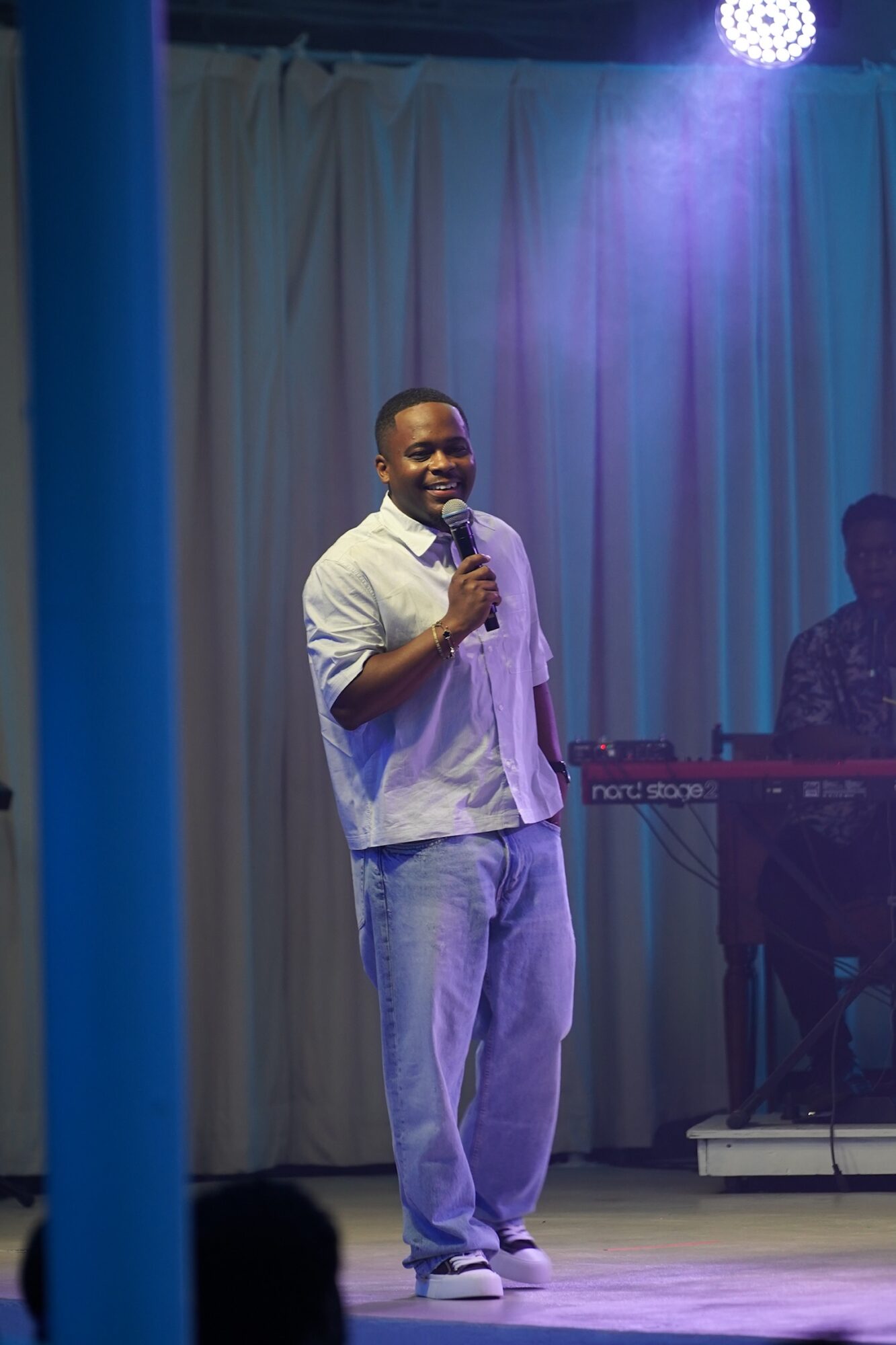 Man holding microphone on stage with curtain backdrop and musician in background.