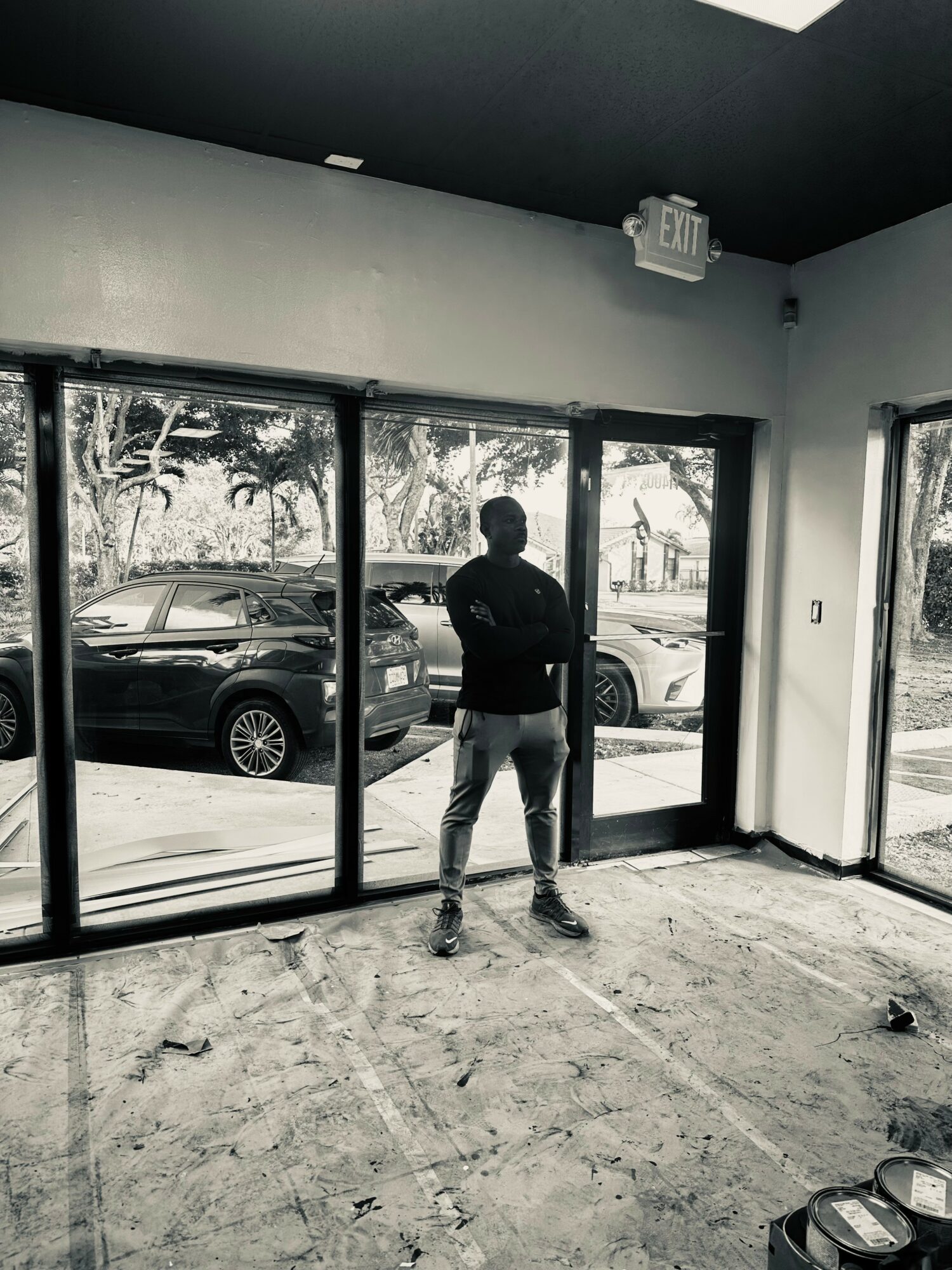 Person standing inside a room with large glass windows and door, looking outside at parked cars and trees.