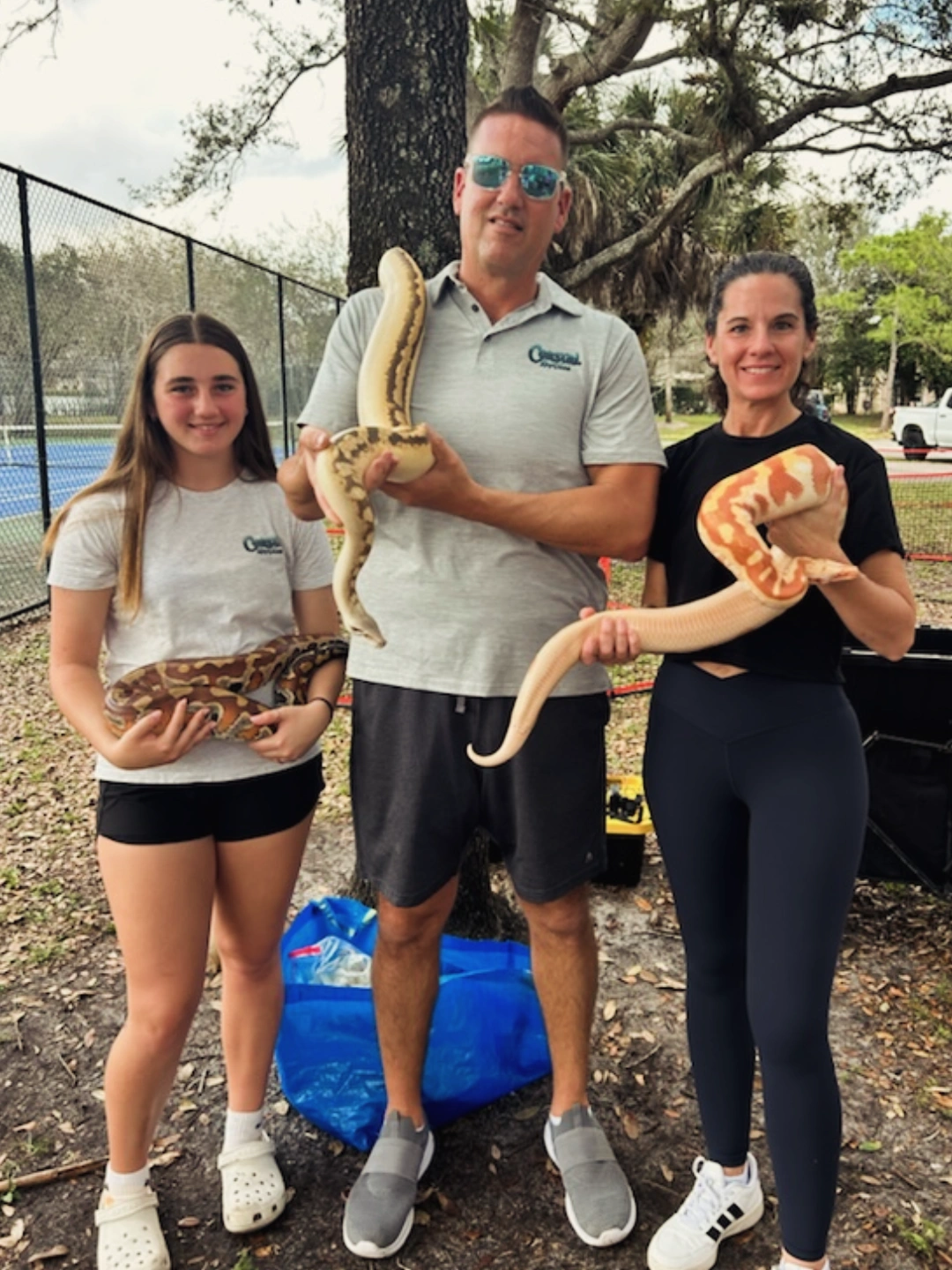 Three people outdoors holding snakes, with trees and a fence in background.