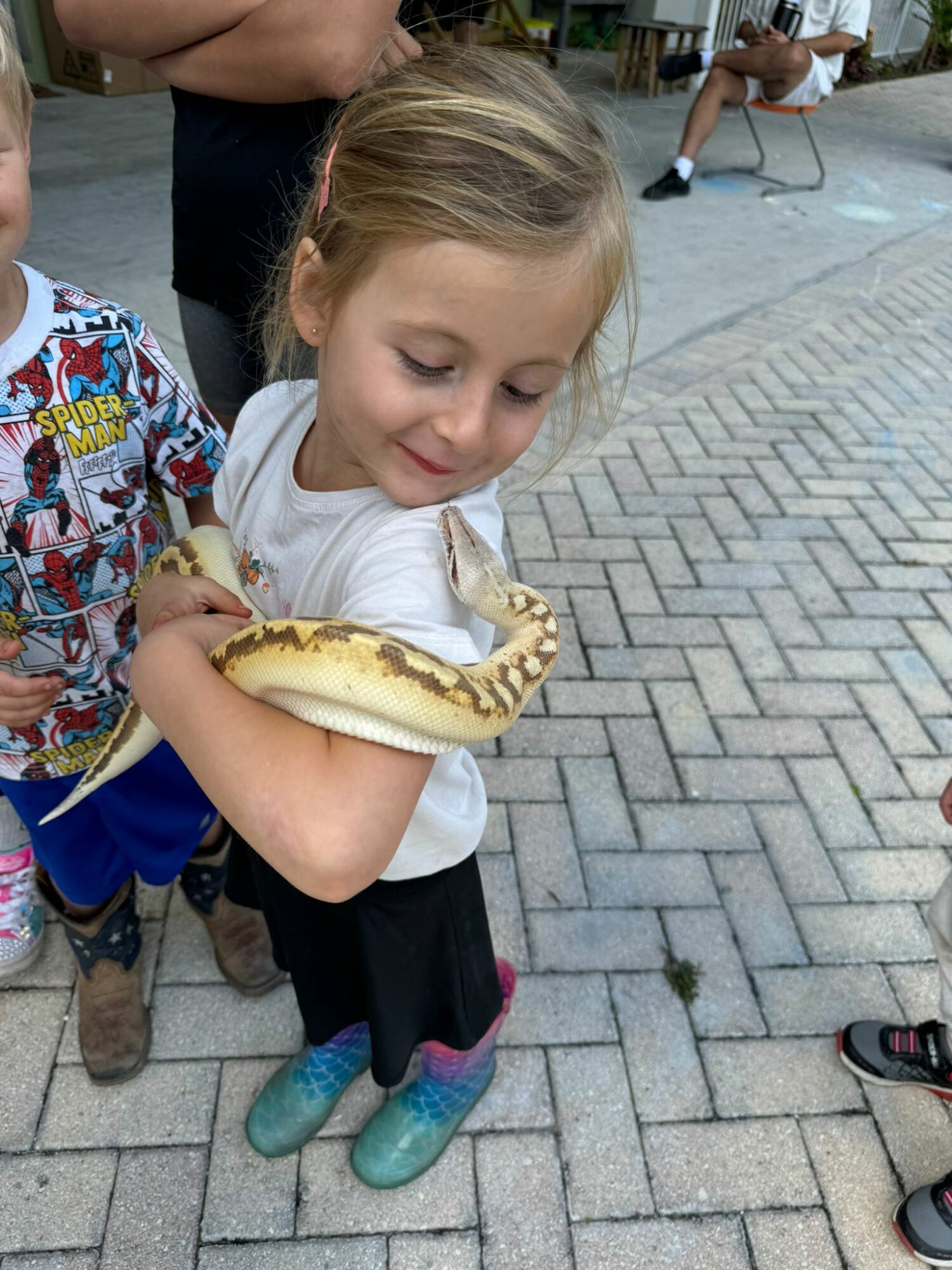 Young girl holding a snake on her arm outdoors, with other children and people in the background.