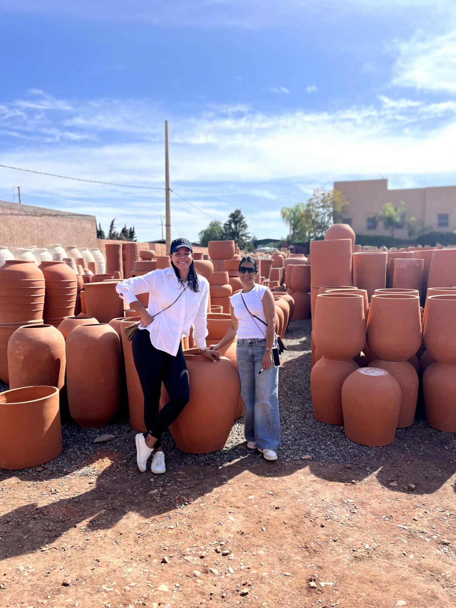 Two people standing among large clay pots outdoors, with a building and blue sky in background.