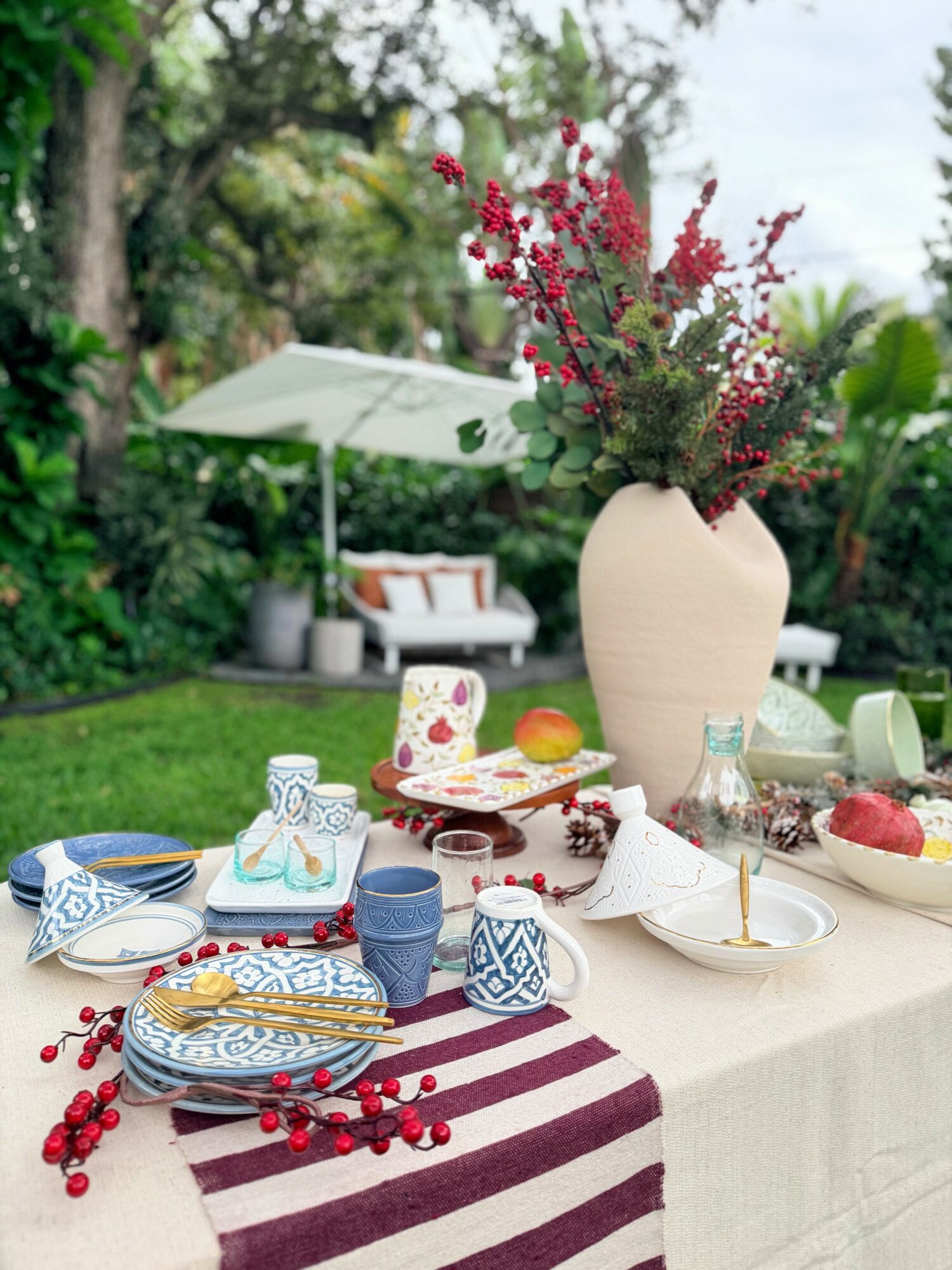 Table with a large vase of flowers, plates, cups, and food, outdoors with greenery and a white umbrella in background.