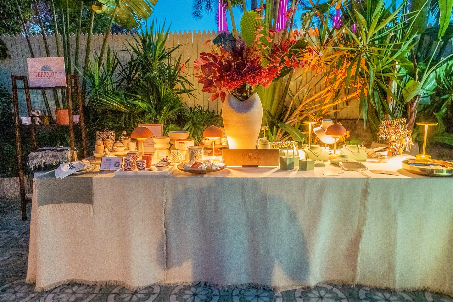 Long table with a white tablecloth, decorated with flowers, candles, and various dishes, outdoors with greenery in background.