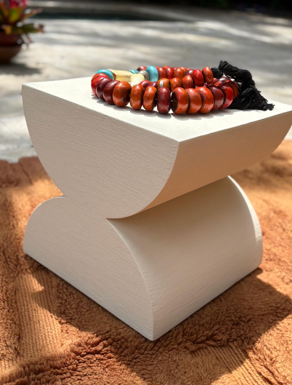 White modern side table with a curved base, outdoors on a brown textured surface, with a beaded necklace on top.