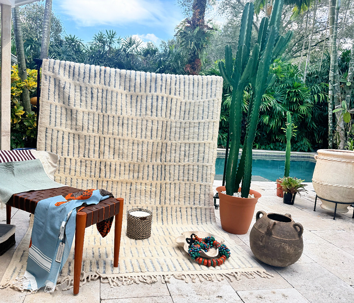 Outdoor patio with large woven wall hanging, potted cactus, pottery, and furniture, overlooking lush greenery and pool.