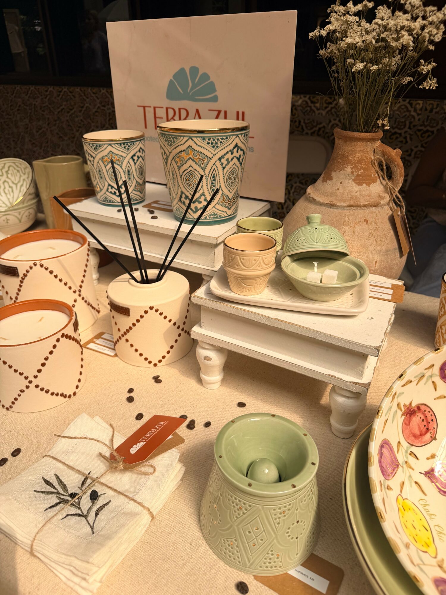 Display of ceramic cups, bowls, and vases with floral and geometric patterns on a table, with a sign and flowers in background.