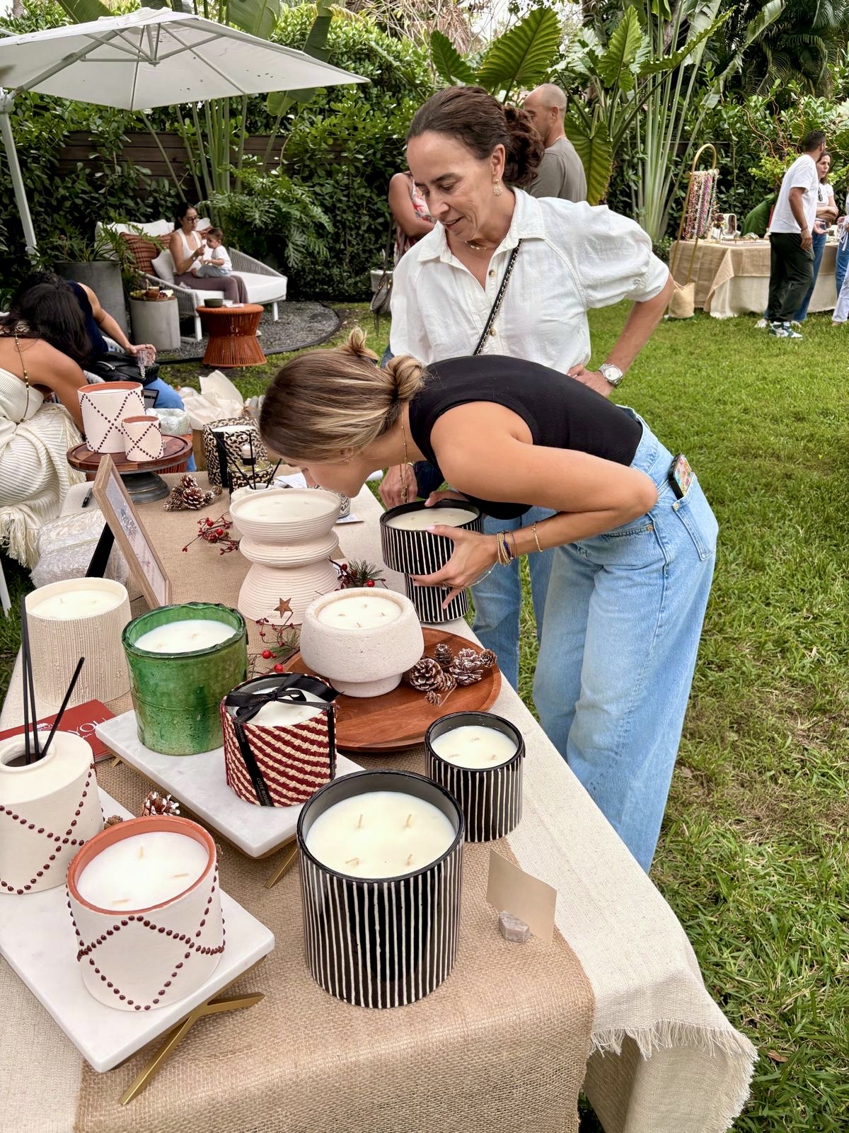 People browsing food and drinks on a table outdoors with greenery and umbrellas in background.