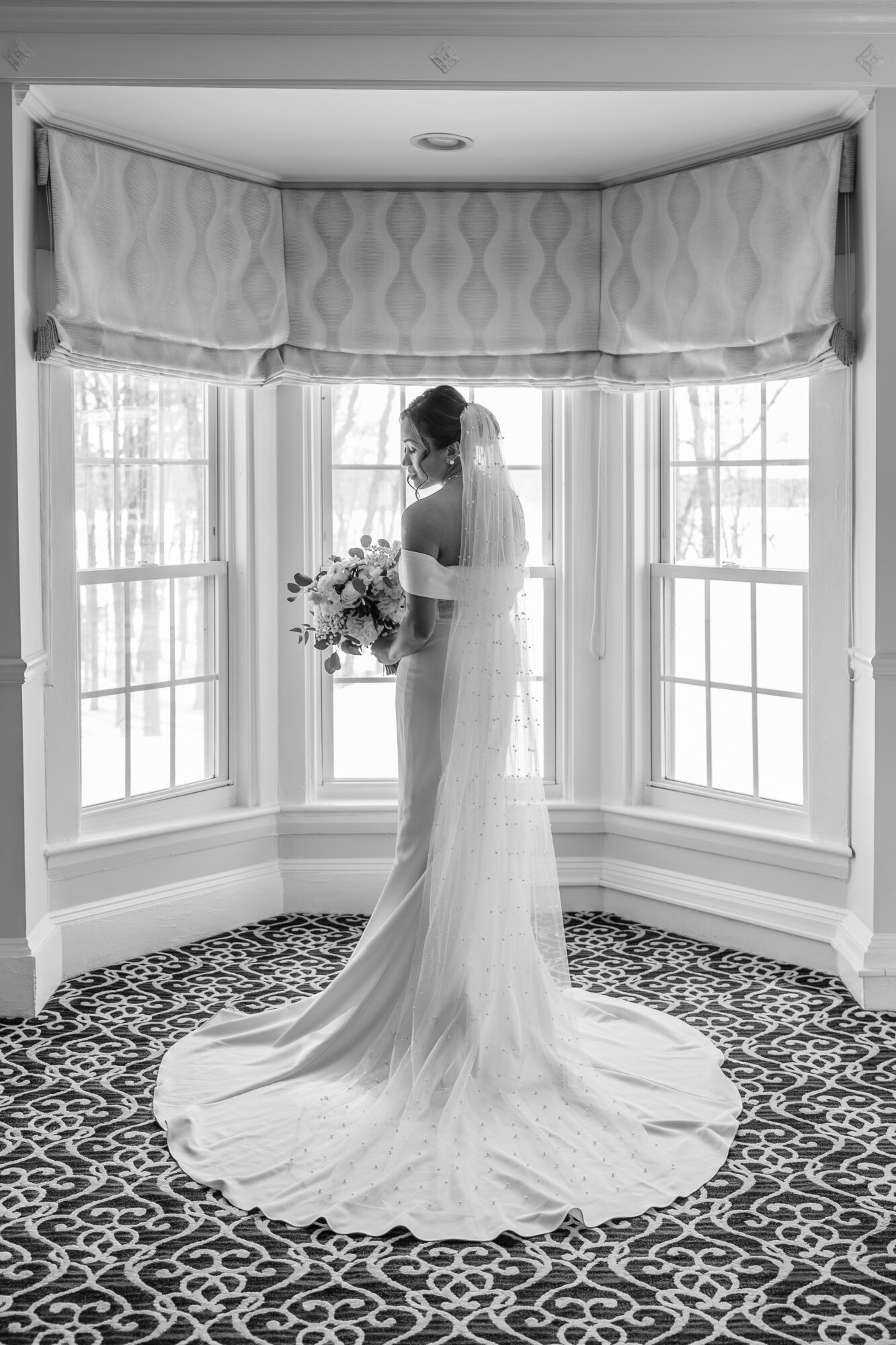 Bride in a long wedding dress holding a bouquet, standing in a bay window with curtains, patterned carpet below.