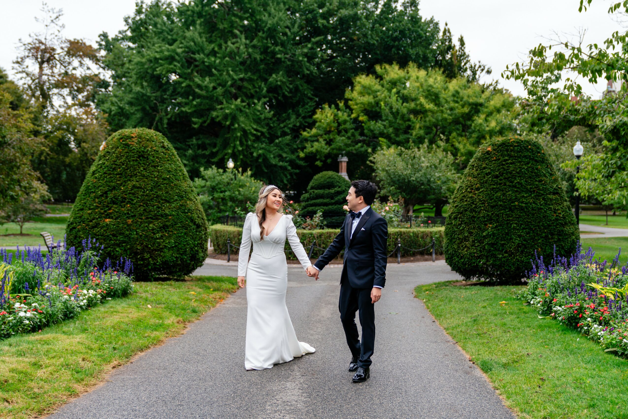 A bride and groom holding hands and walking on a garden path surrounded by greenery and flowers.