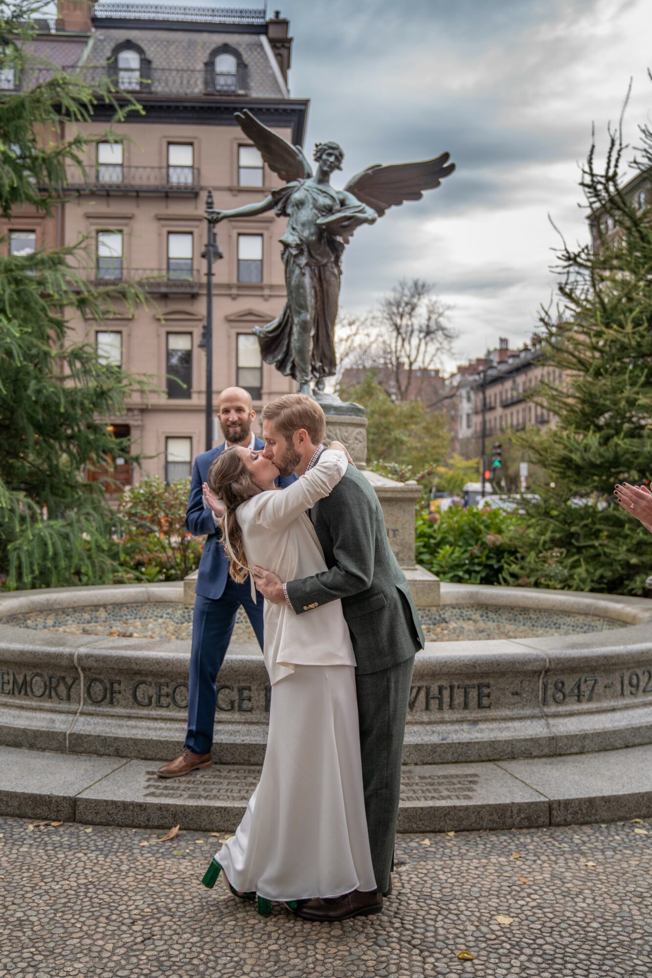 Couple embracing in front of a statue of an angel with wings, surrounded by trees and buildings.