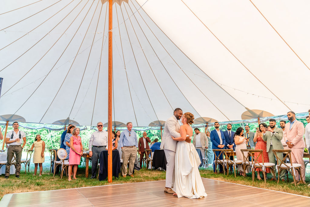 Couple dancing under a large tent with guests standing and sitting nearby outdoors.
