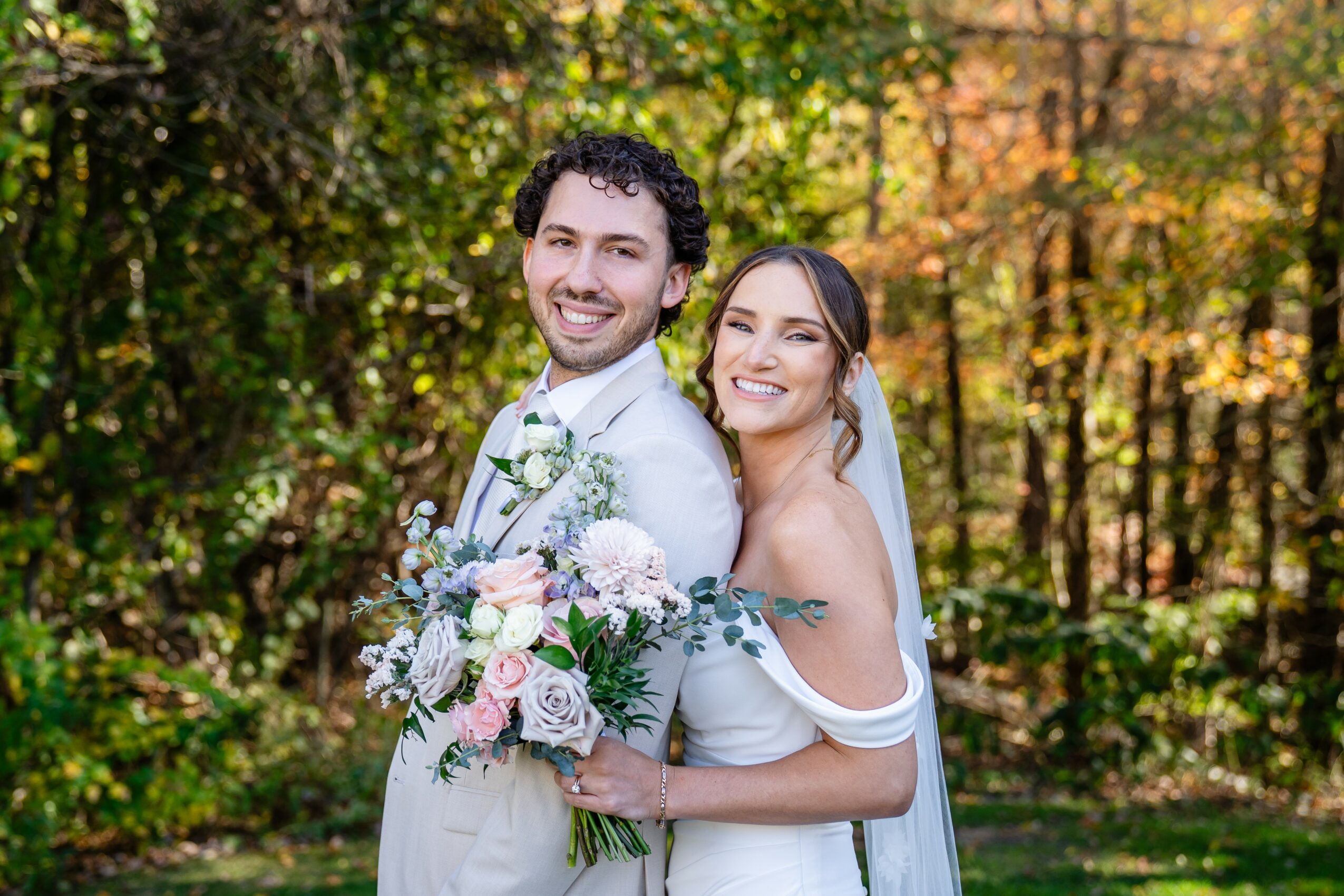 Happy couple outdoors with trees and autumn leaves in background, holding a bouquet of flowers.