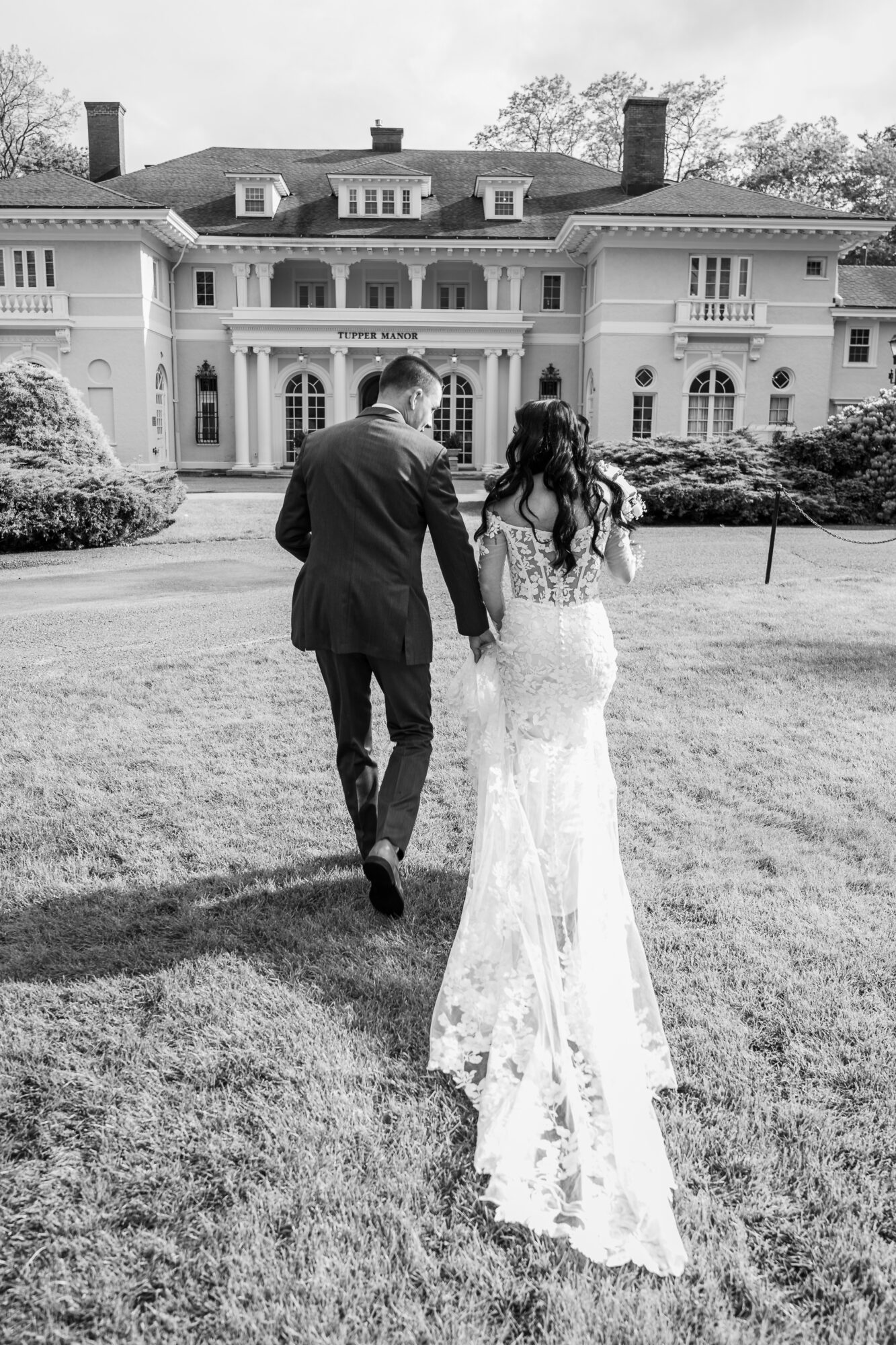 Couple walking towards a large mansion, holding hands, with a lawn and bushes in front.