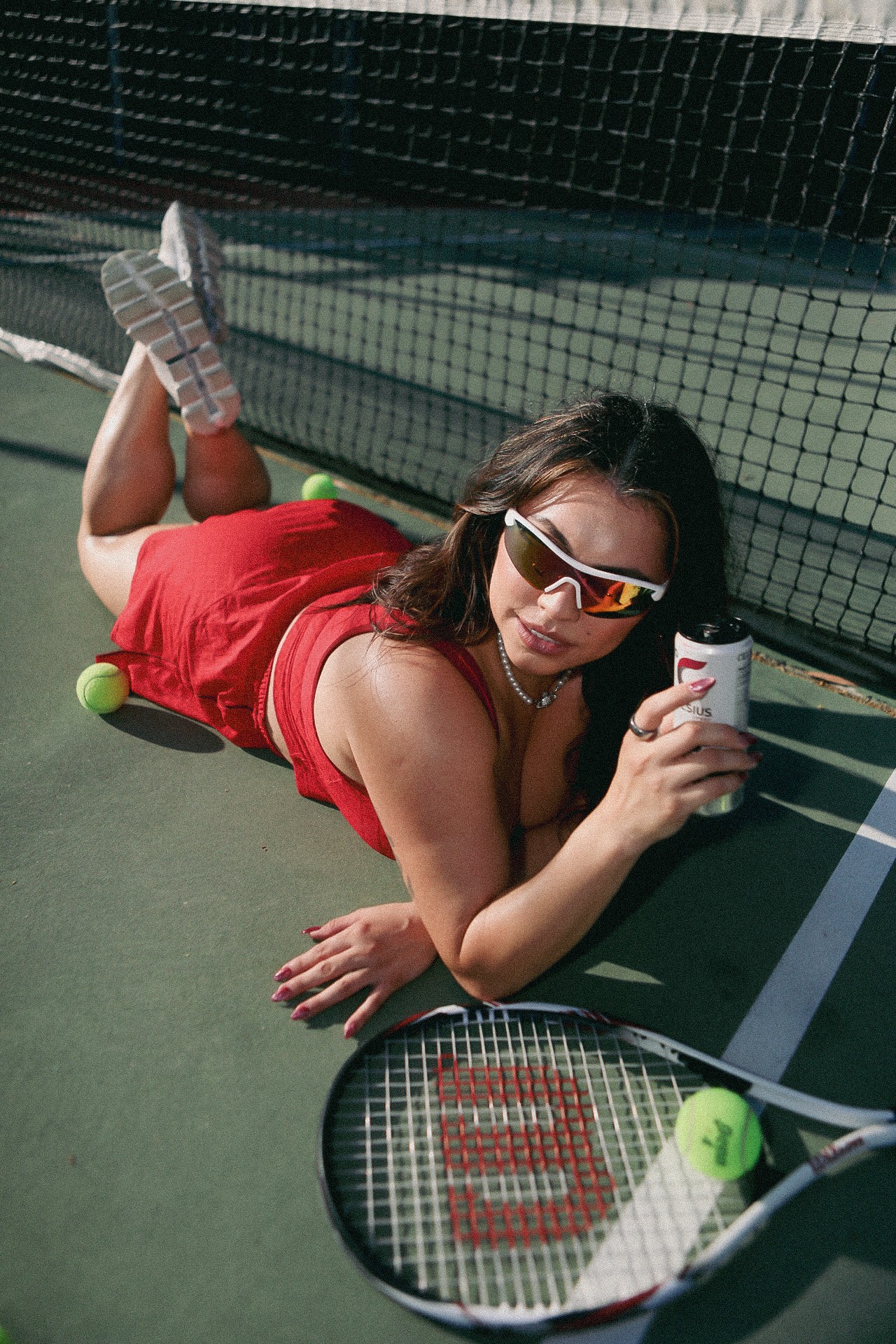 Woman in red dress and sunglasses lying on tennis court holding a smartphone, tennis racket and balls nearby.