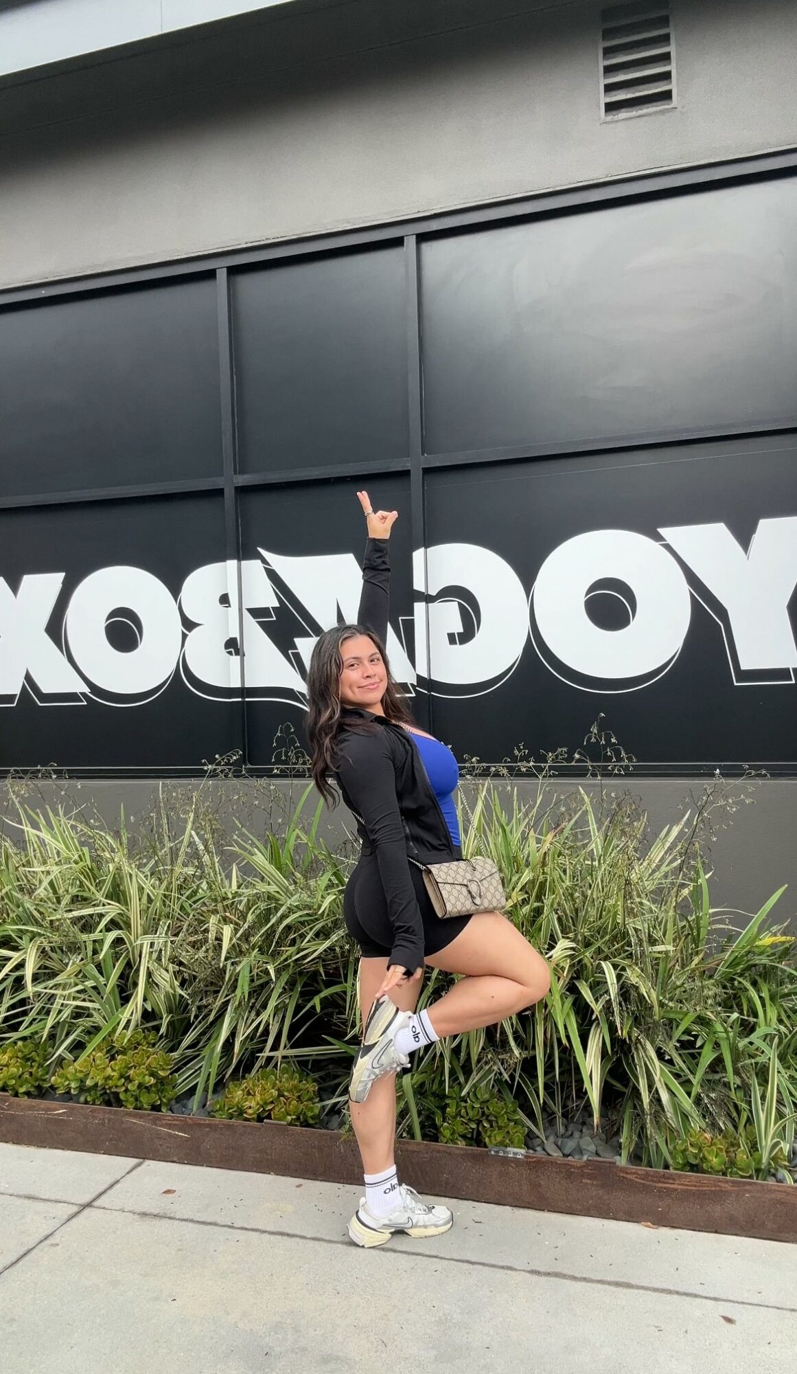 Young woman standing outdoors near a large black wall with white text, pointing upward with one hand, surrounded by green plants.
