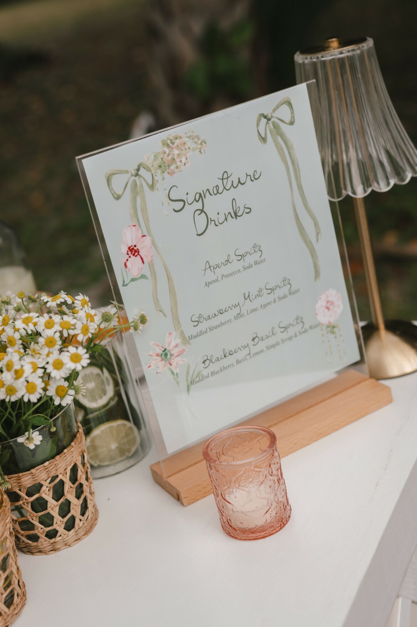 Signage with floral decorations and handwritten text, placed on a table with flowers, a glass, and a lamp.