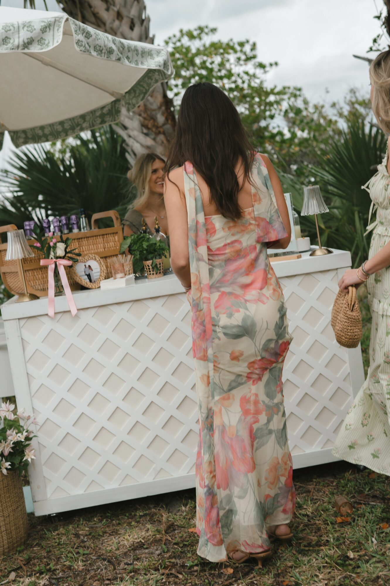 Woman with long dark hair in floral dress standing near a white lattice table outdoors, surrounded by greenery and people.