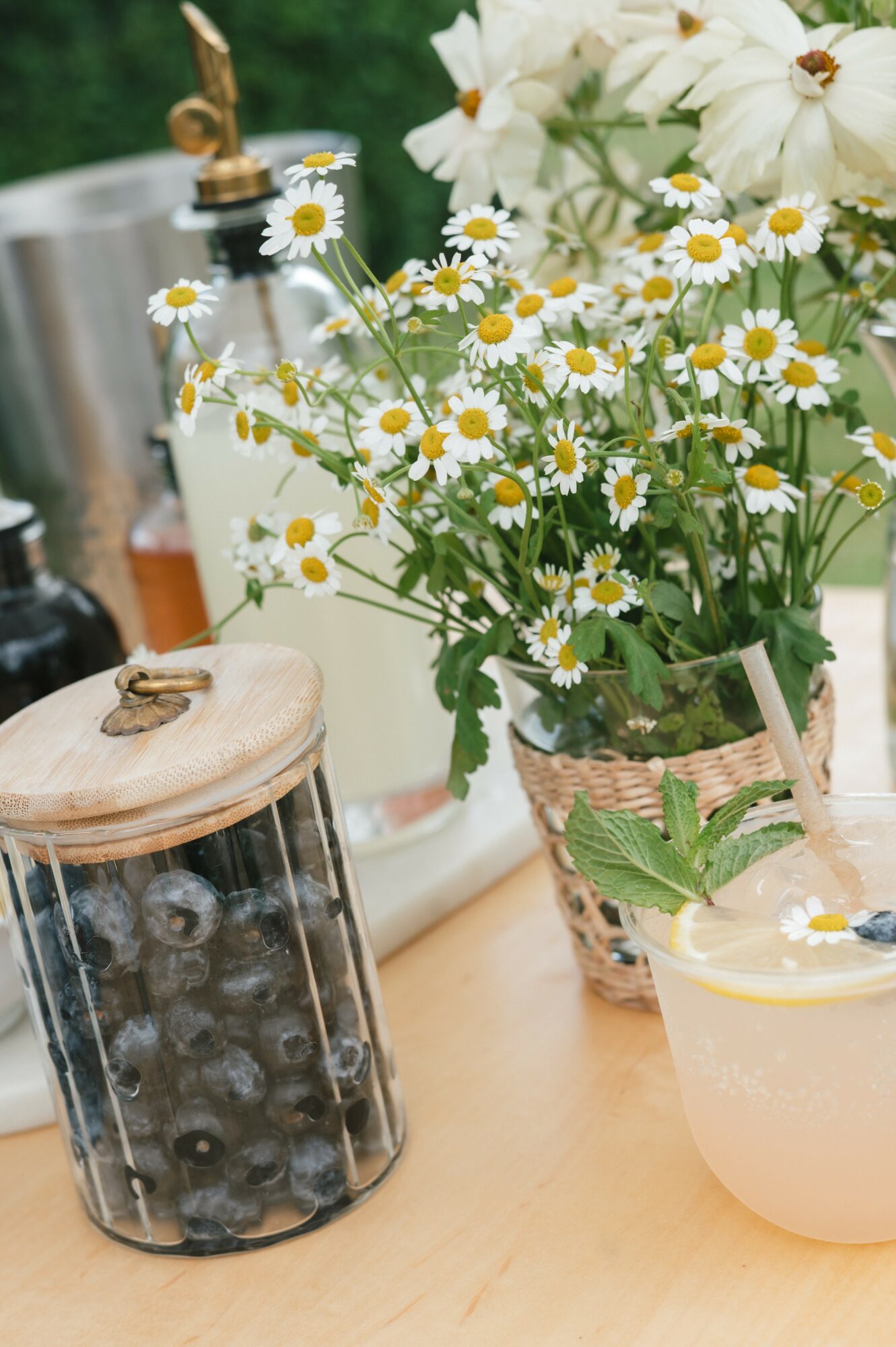 Vase with daisies, jar of blueberries, and a candle on a wooden surface.