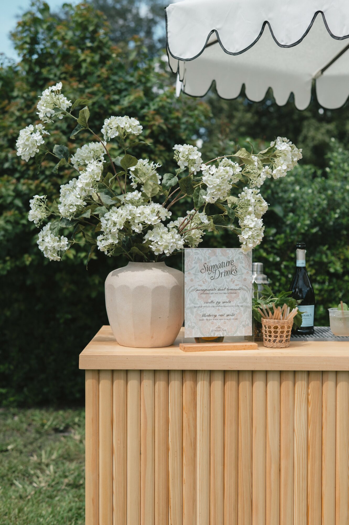 Vase with white flowers on wooden table outdoors, umbrella overhead, bottle, basket, and card present.