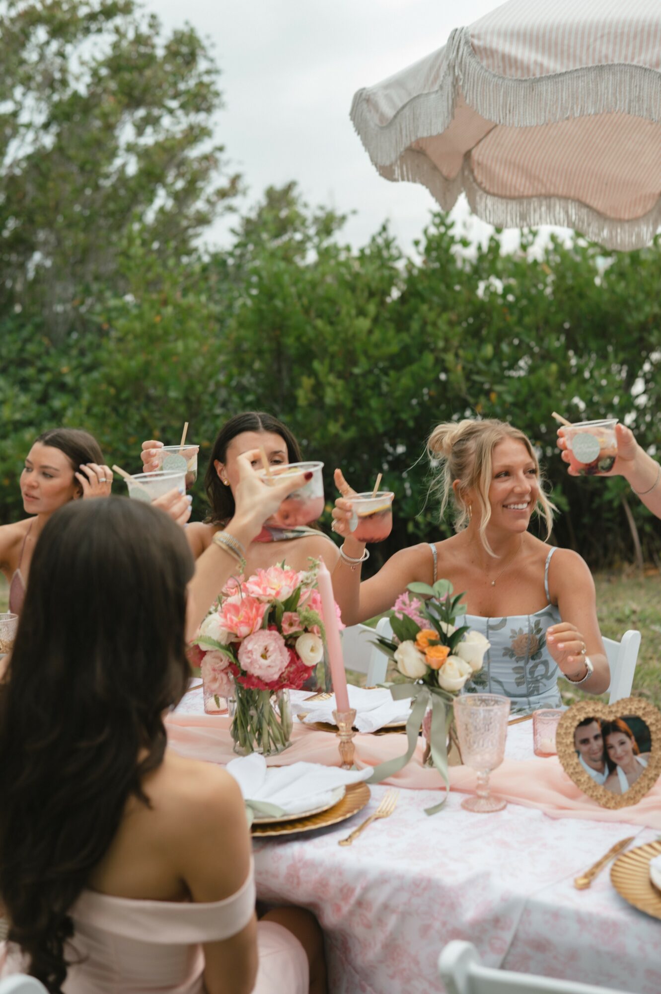 Group of women celebrating outdoors with drinks and floral arrangements on a table, smiling and raising glasses.