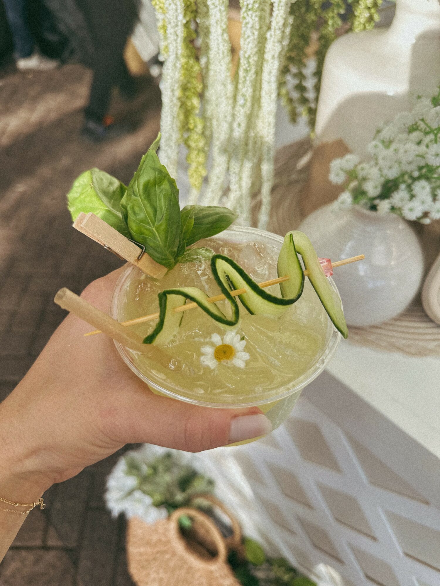 Hand holding a cocktail garnished with basil, cucumber slices, and a green spiral straw, with a blurred floral background.